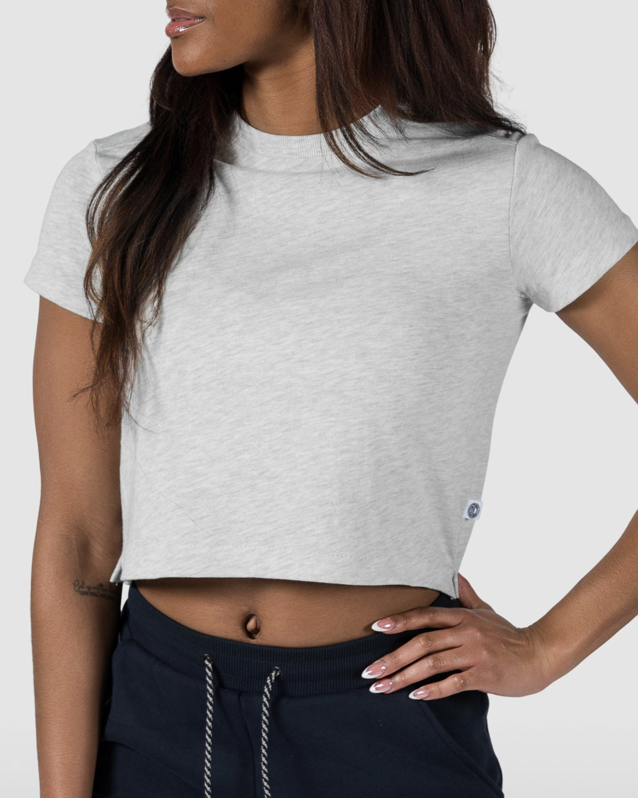 A woman in the BEARDEDGOAT Womens Valley Tee, a light gray cropped shirt, and dark jogger pants stands with one hand on her hip against a plain background—a stylish, casual summer look.