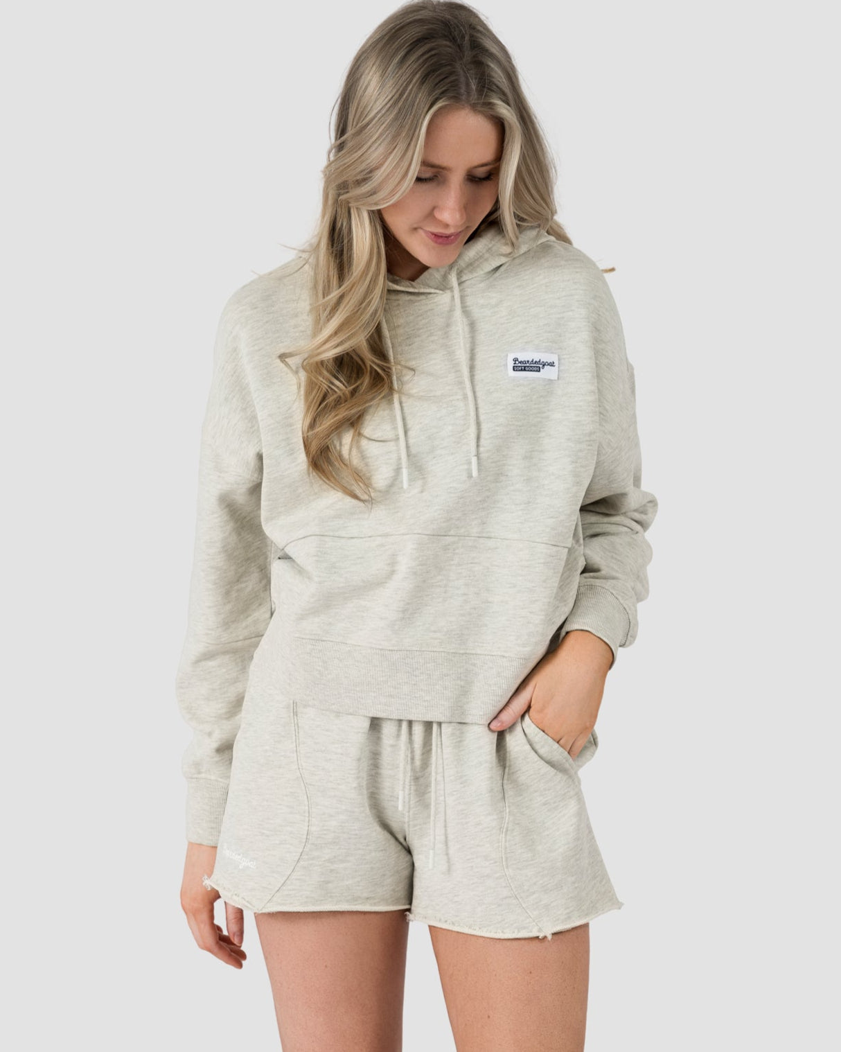 A person stands against a plain background in BEARDEDGOAT’s light gray hooded sweatshirt and relaxed-fit Womens Drift Short with white sneakers. One hand is in their pocket as they look down—a laid-back summer staple look.