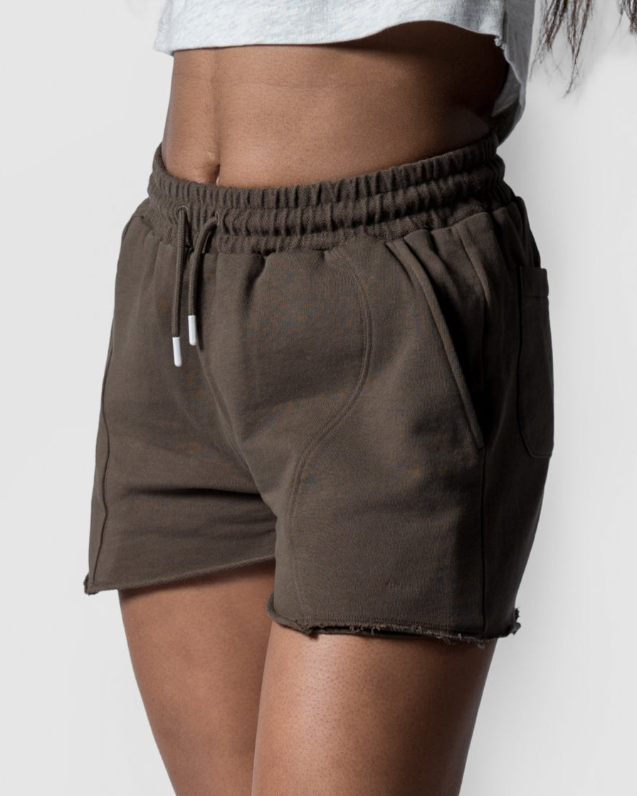 A person stands against a plain light gray background wearing BEARDEDGOATs Womens Drift Short in dark olive green and white sneakers, with a light gray crop top—showcasing this relaxed fit summer staple from the waist down.