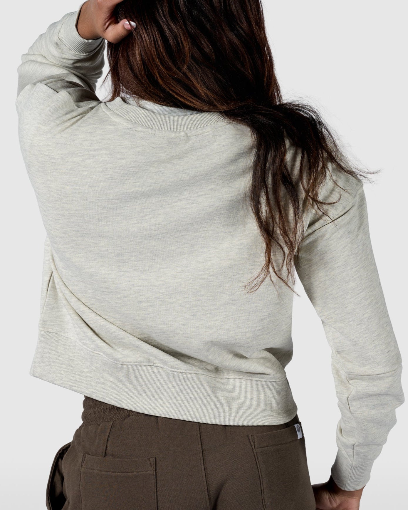 A person faces away, wearing BEARDEDGOATs Womens Drift Crew sweatshirt in light gray with brown shorts. One hand touches the back of their head against a plain, light background.