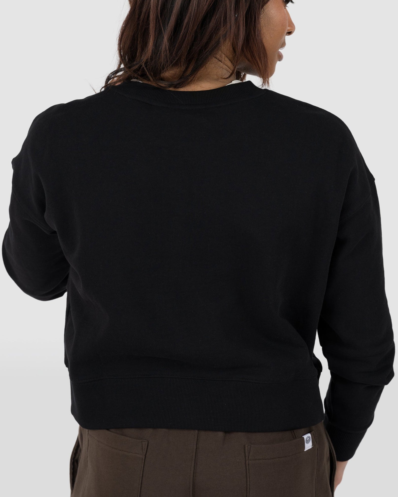 A person with long, dark hair is seen from behind wearing the BEARDEDGOAT Womens Drift Crew sweatshirt and brown pants, standing in front of a plain light gray background.