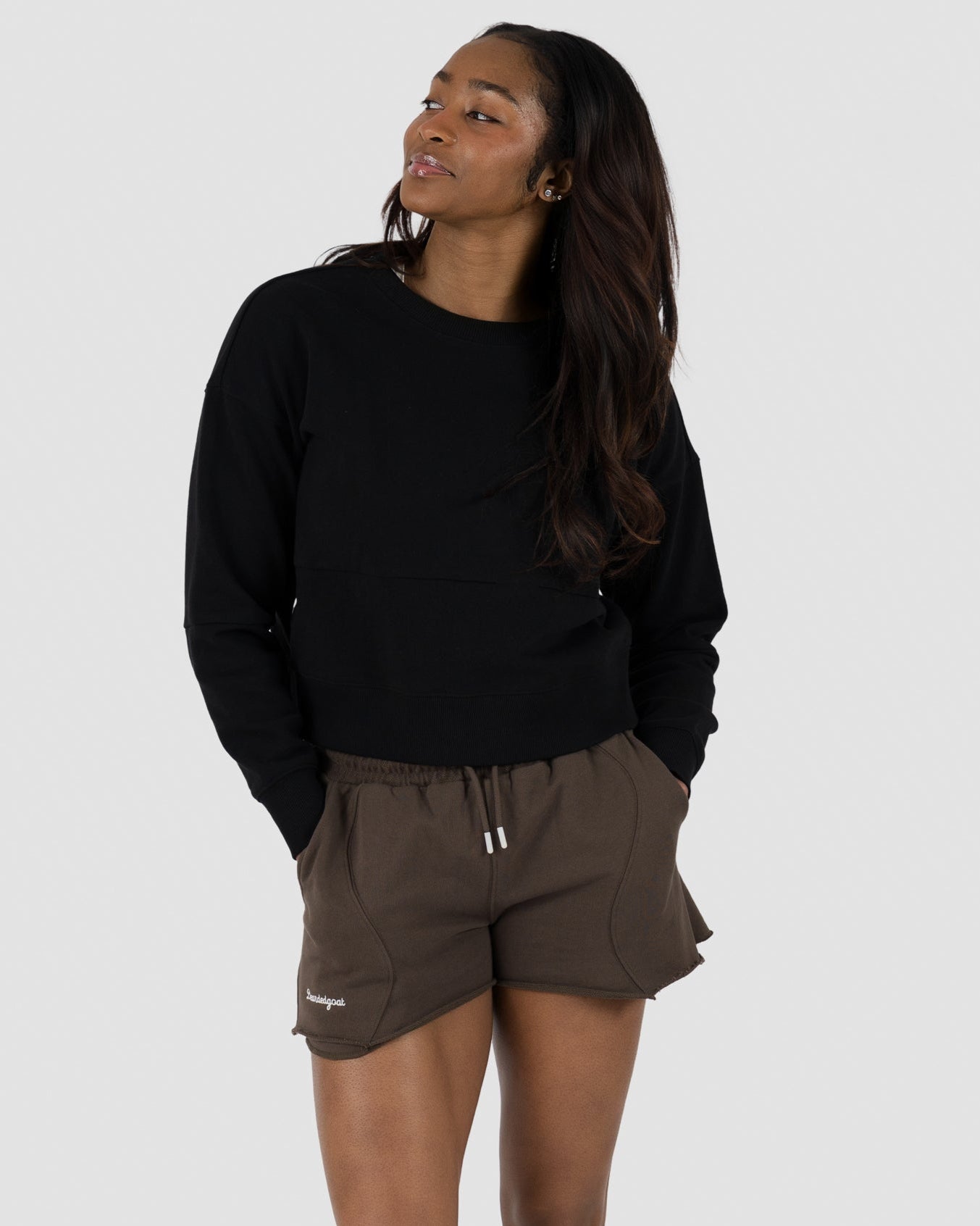A woman stands against a plain background, smiling to the side. She wears the BEARDEDGOAT Womens Drift Crew sweatshirt, brown drawstring shorts, and white sneakers, with her hands in her pockets.