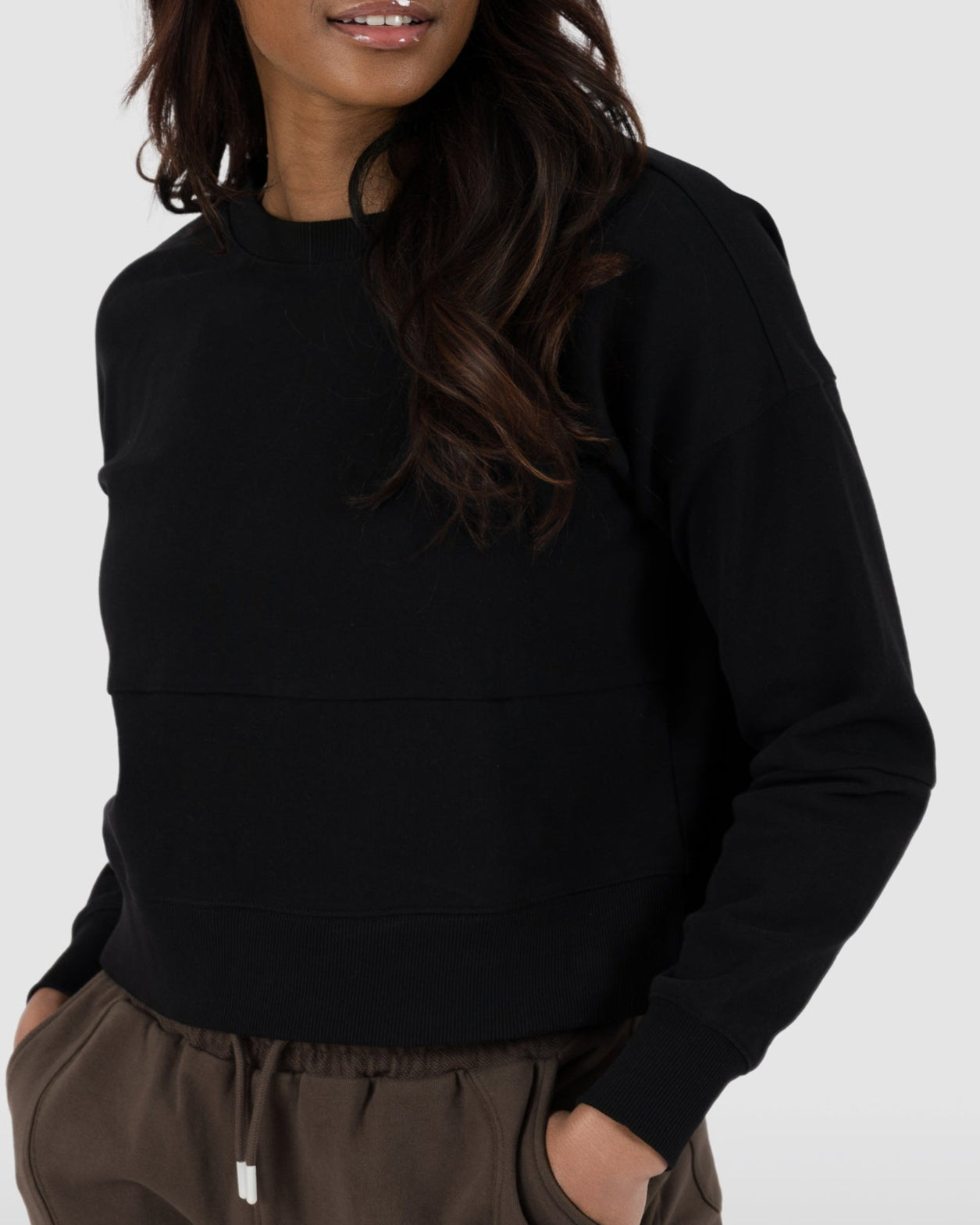 A woman with long dark hair smiles to the side with her hands in her pockets, modeling the BEARDEDGOAT Womens Drift Crew sweatshirt against a plain light background.