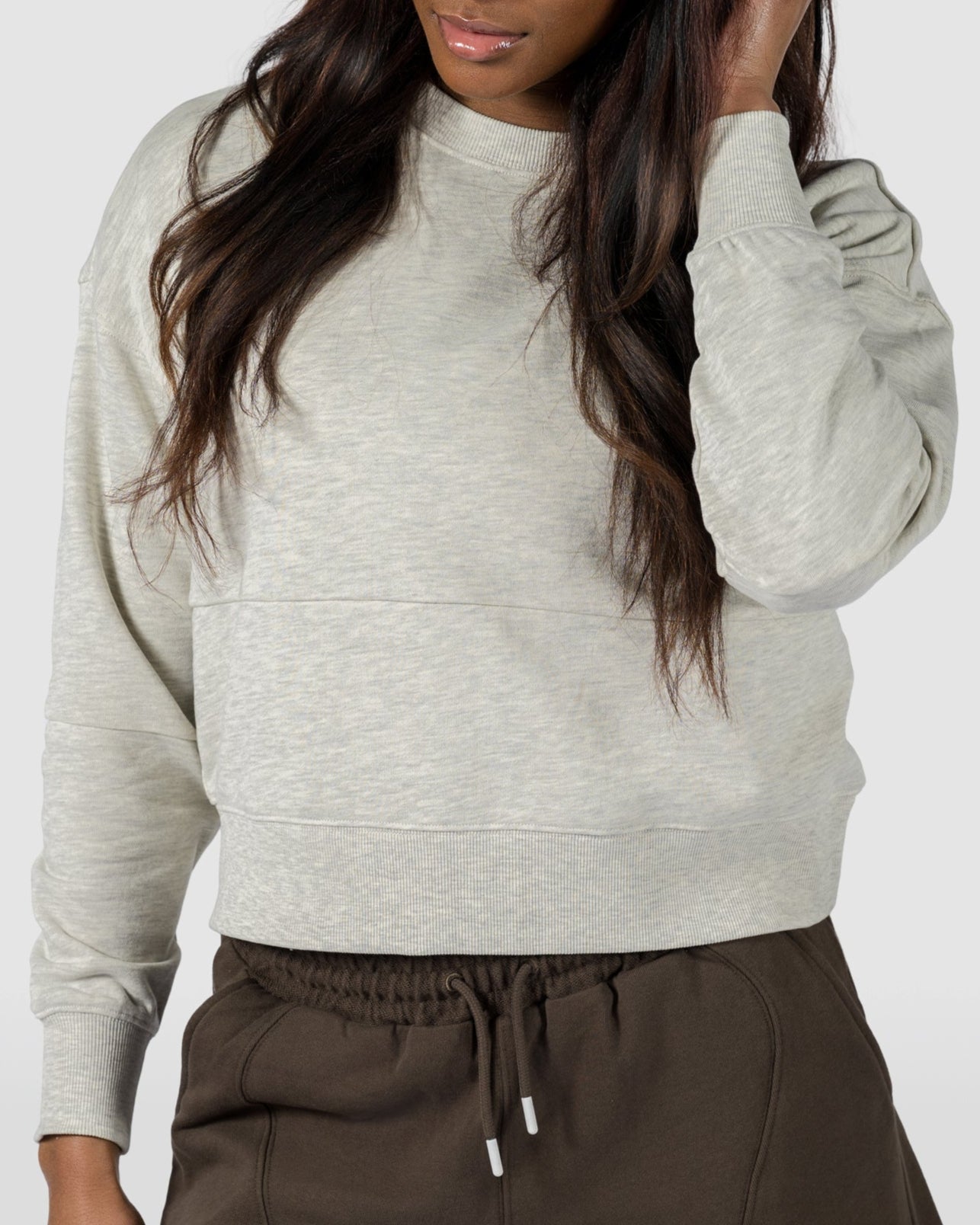 A woman stands against a plain light background, wearing the BEARDEDGOAT Womens Drift Crew in soft terry and dark brown shorts. She looks down, touching her hair with one hand and maintaining a neutral expression.