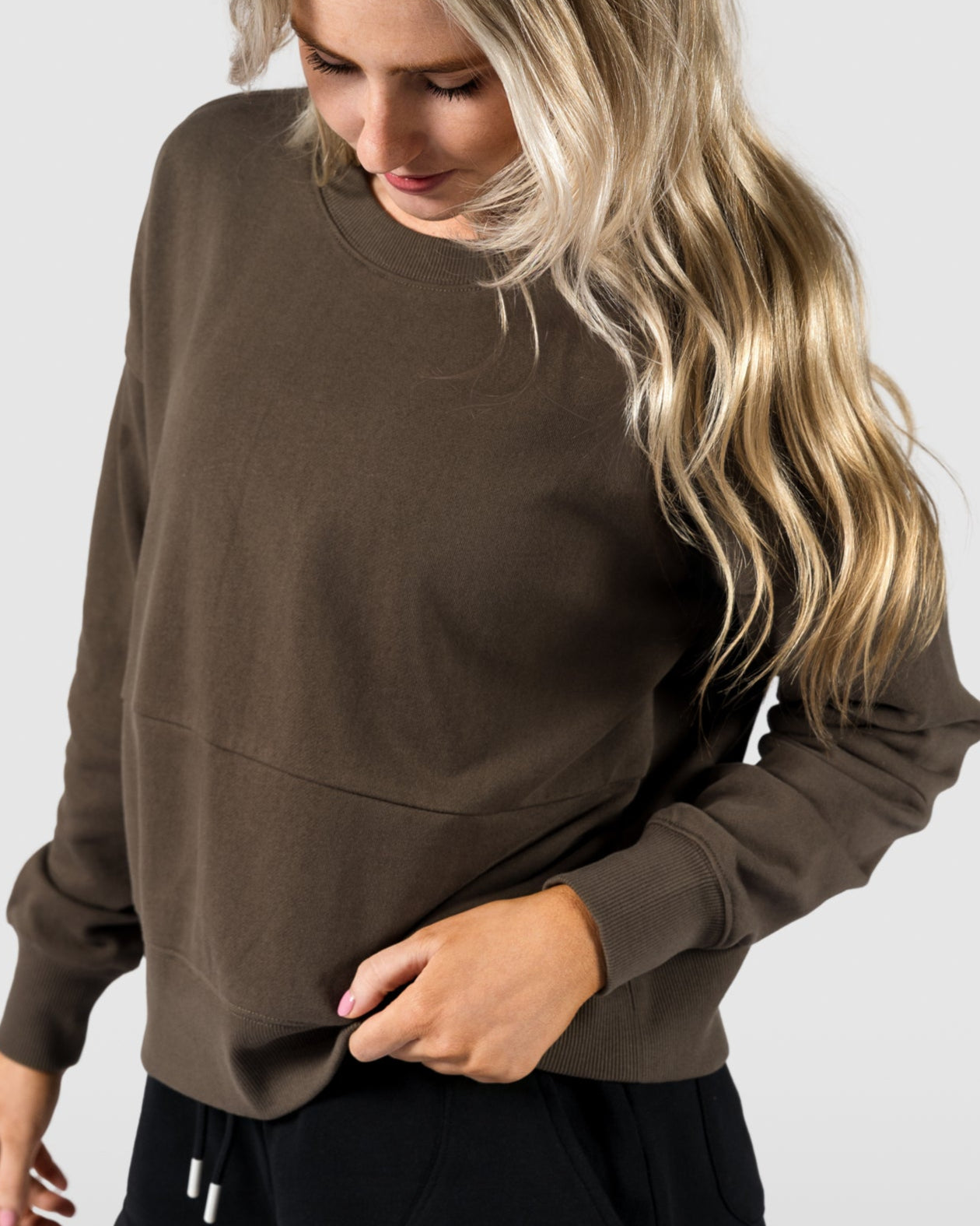 A woman with long blonde hair wears the BEARDEDGOAT Womens Drift Crew in soft terry fabric and black shorts, standing against a plain light gray background while looking down and touching her sweatshirt.