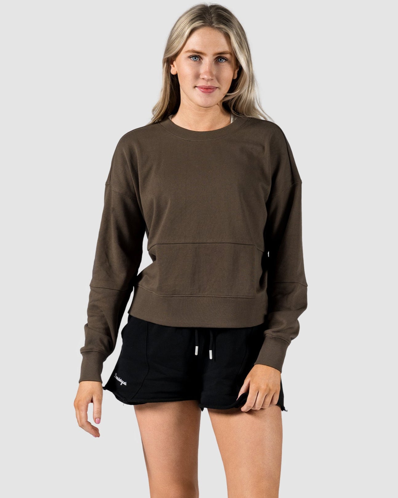 A woman faces forward against a light gray background, wearing the BEARDEDGOAT Womens Drift Crew sweatshirt in soft terry fabric, paired with black drawstring shorts and white sneakers.