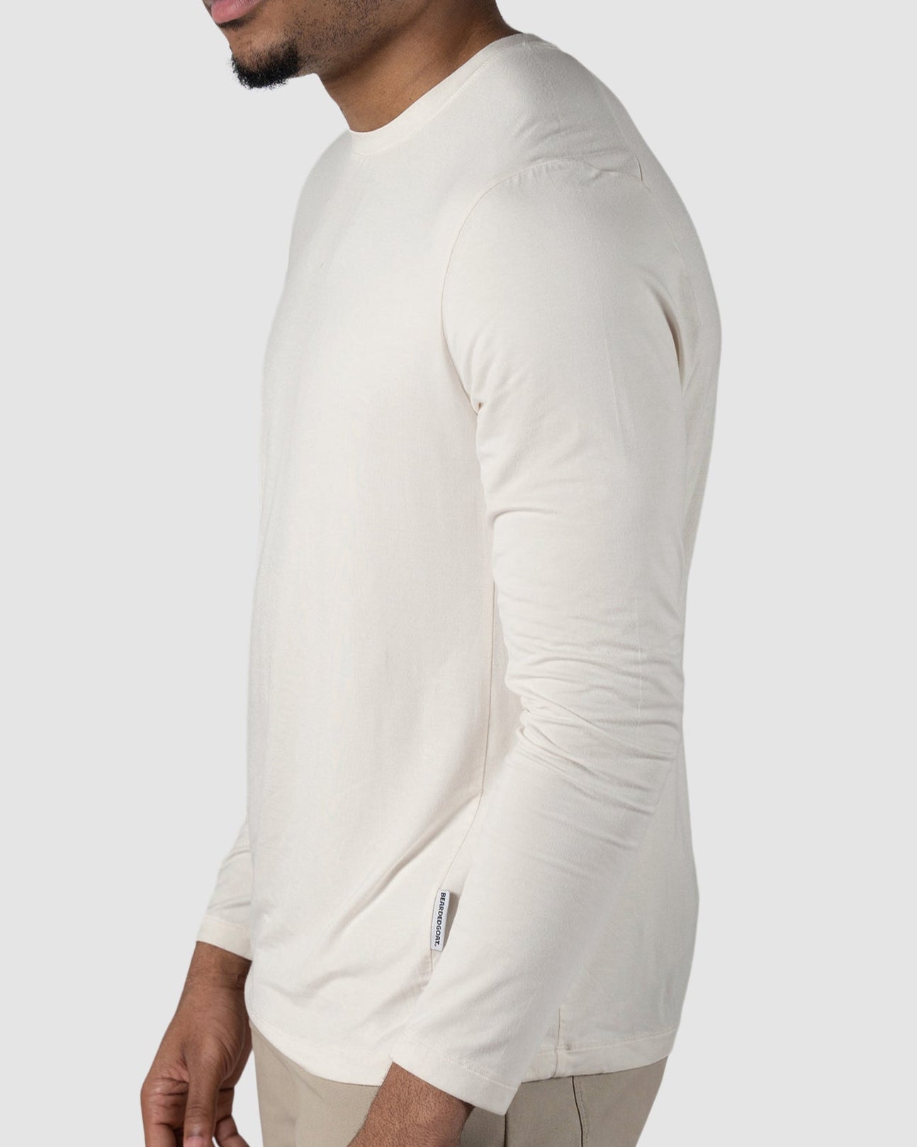 A man wears the BEARDEDGOAT Mens EverZen Long Sleeve in off-white and beige pants, standing against a light gray background, looking slightly left with a neutral expression.