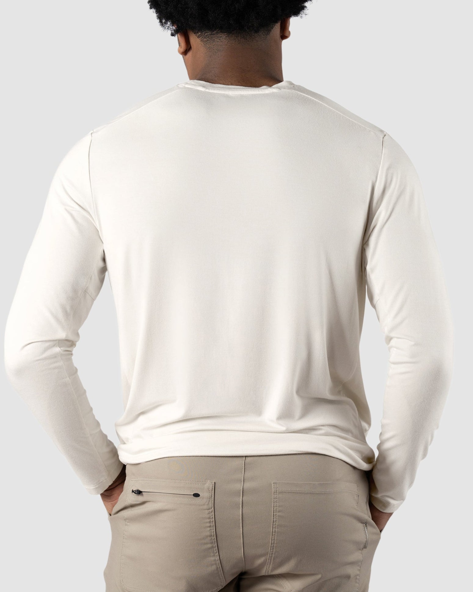 A man with short, curly hair is seen from behind, wearing the BEARDEDGOAT Mens EverZen Long Sleeve shirt in off-white and light beige pants, standing against a plain light gray background.