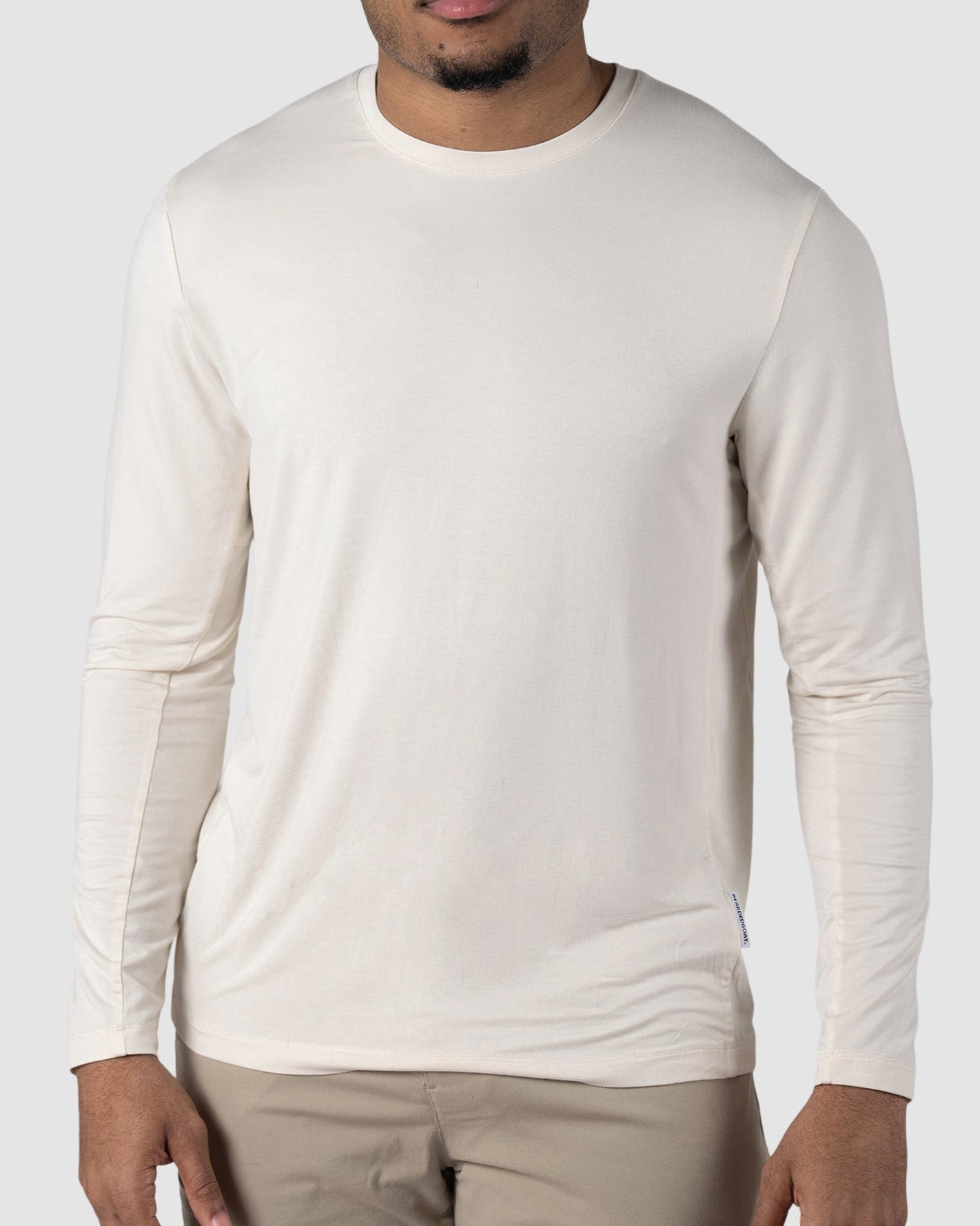 A man with short curly hair, dressed in the BEARDEDGOAT Mens EverZen Long Sleeve—a long-sleeve, off-white, odor-resistant bamboo fiber shirt—and beige pants stands facing the camera against a plain light gray background.