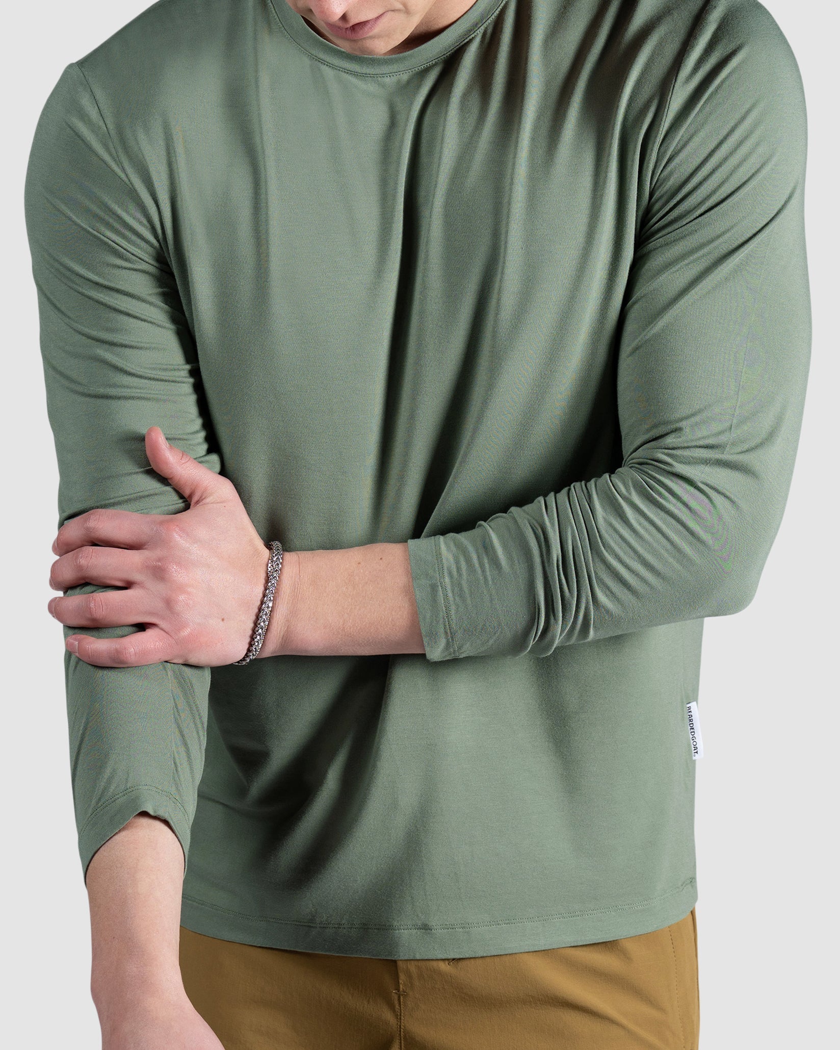 A man wearing the BEARDEDGOAT Mens EverZen Long Sleeve in light green and tan shorts stands against a plain background, adjusting his left sleeve with his right hand.