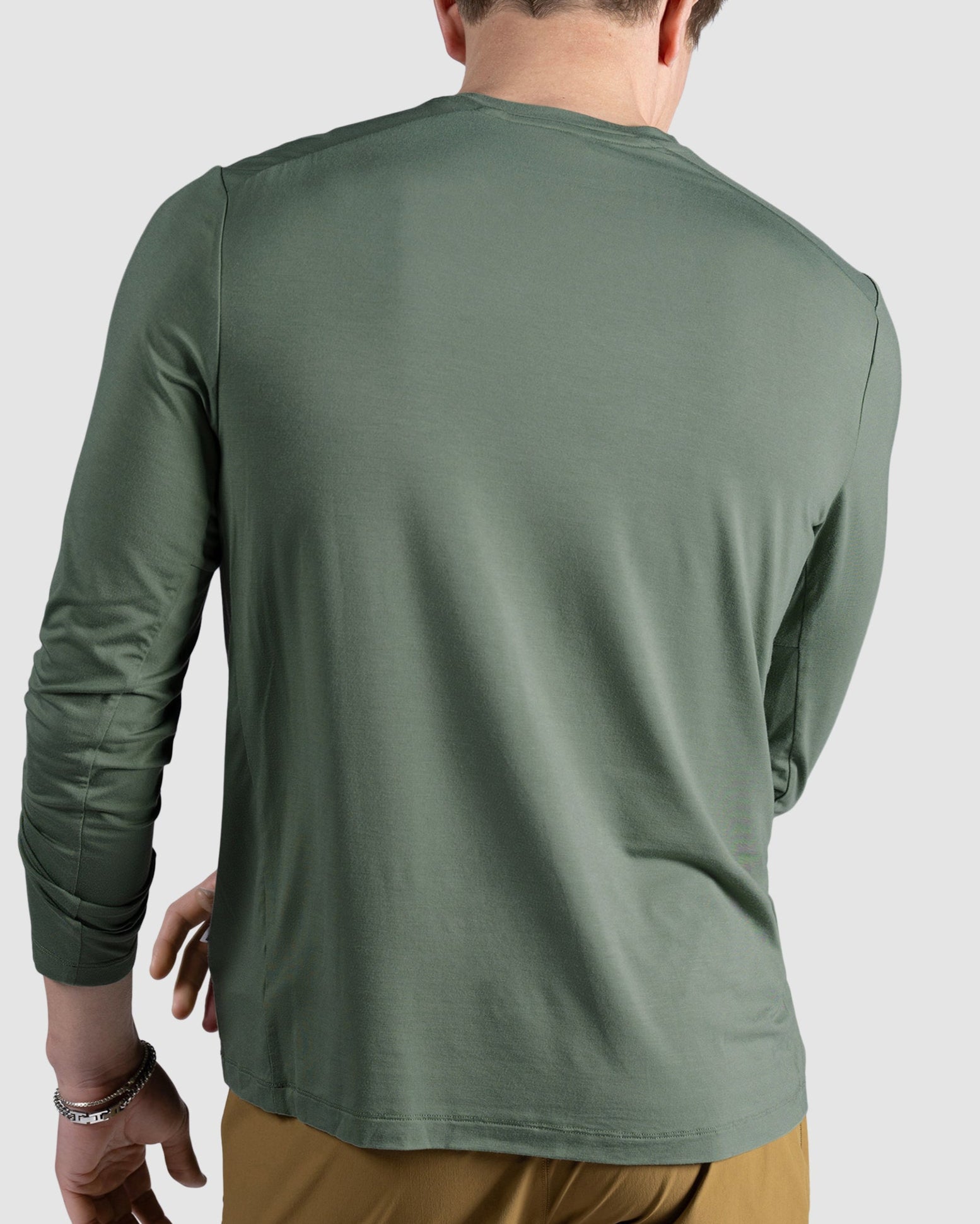 A person faces away from the camera wearing the BEARDEDGOAT Mens EverZen Long Sleeve in green and tan pants, with their left arm bent and head bowed slightly forward against a plain light background.