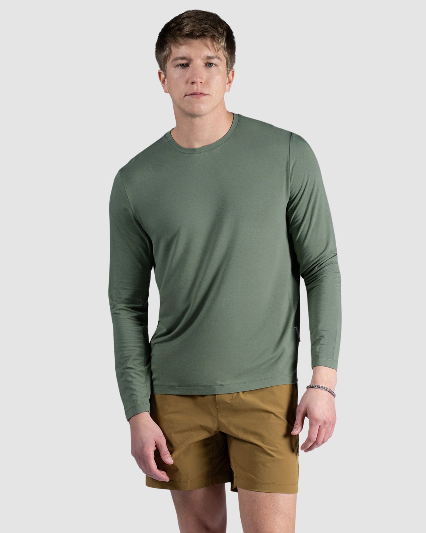 A young man with short light brown hair and a neutral expression stands against a light gray background wearing the BEARDEDGOAT Mens EverZen Long Sleeve in olive green, paired with brown shorts, white socks, and white sneakers.