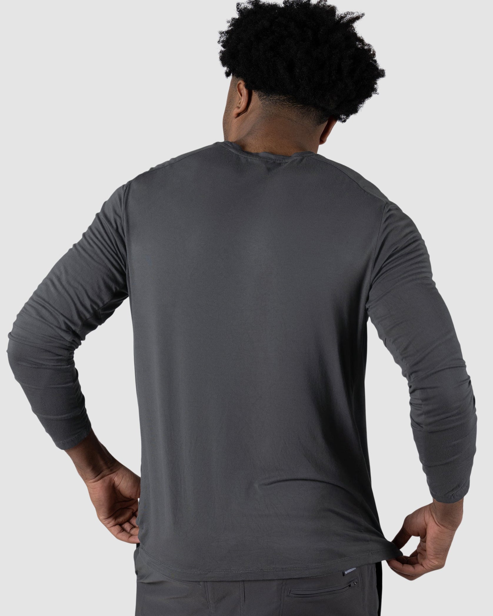 A man with short curly hair is seen from behind, wearing the BEARDEDGOAT Mens EverZen Long Sleeve and matching gray pants made of moisture-wicking fabric, standing against a plain light gray background.