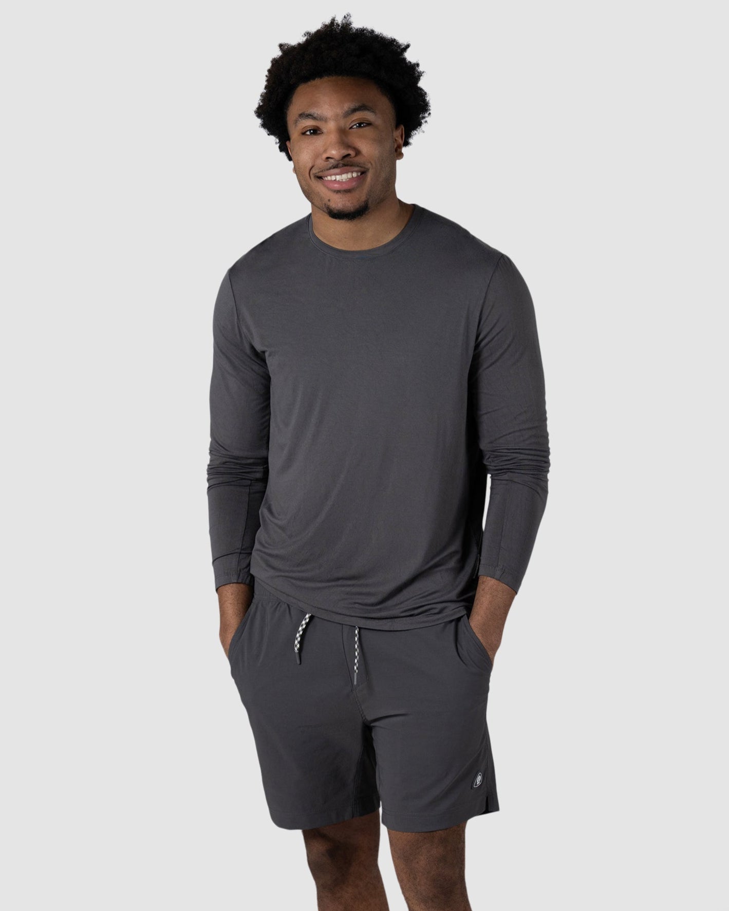 A man smiles with his hands in his pockets, wearing the BEARDEDGOAT Mens EverZen Long Sleeve in dark gray, gray shorts, and white sneakers against a plain light gray background.