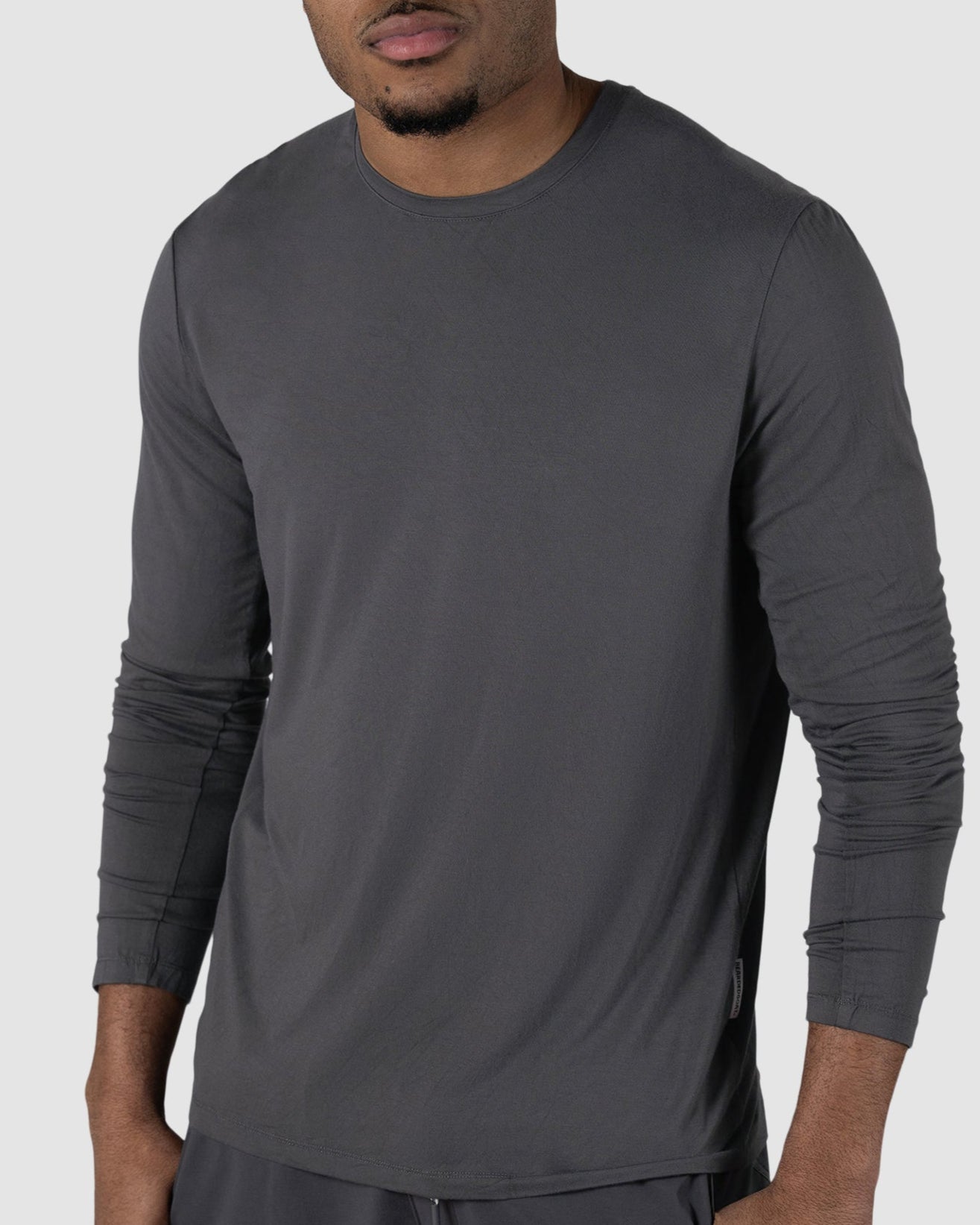 A man with short curly hair wears the BEARDEDGOAT Mens EverZen Long Sleeve in dark gray and dark pants, standing against a light gray background. He looks at the camera with a neutral expression. The shirt is odor-resistant and made from bamboo fibers.