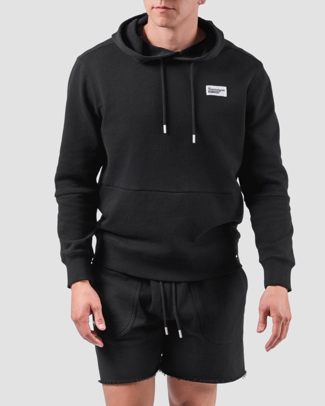 A man wears the BEARDEDGOAT Mens Drift Hoodie in black, paired with matching terry cloth shorts, white crew socks, and white sneakers against a light gray background—ideal for a spring-to-summer look.