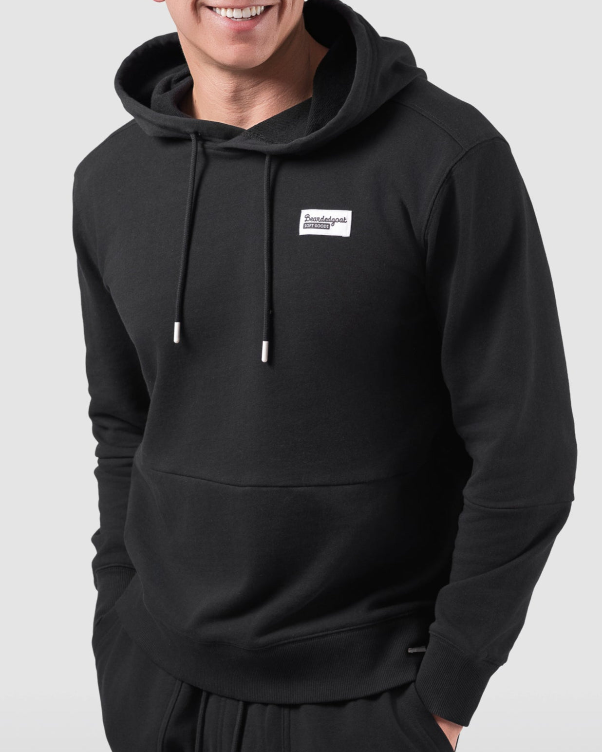 A young man smiles while wearing the BEARDEDGOAT Mens Drift Hoodie, paired with matching black shorts, standing against a plain light gray background.