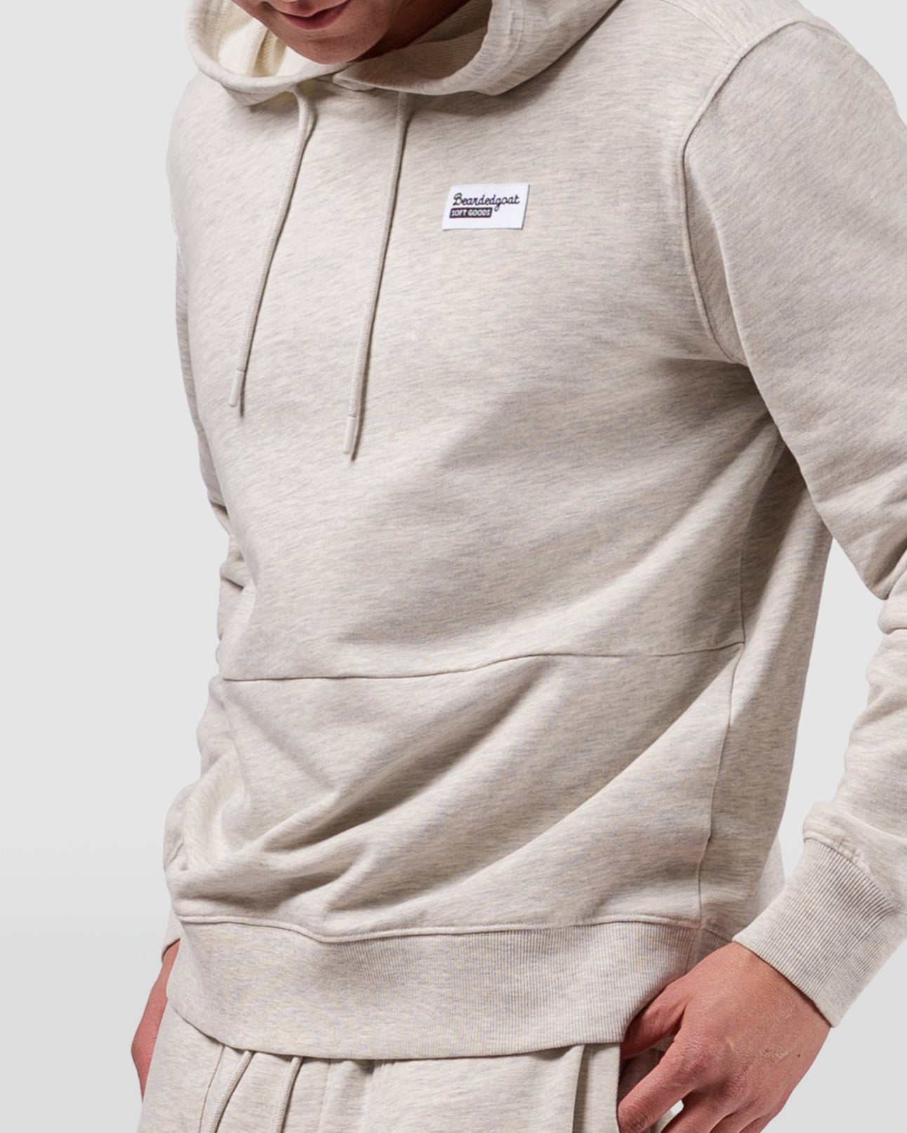 A young man wears the BEARDEDGOAT Mens Drift Hoodie in light beige terry cloth with a small rectangular chest logo and matching sweatpants, one hand in his pocket, against a plain light gray background.