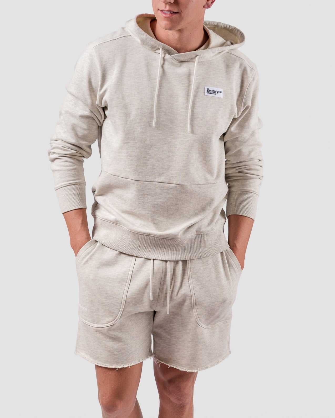 A young man stands against a light gray background, wearing the BEARDEDGOAT Mens Drift Hoodie and matching beige terry cloth shorts—perfect for spring to summer. He has short hair, looks to the side, and keeps his hands in his pockets.