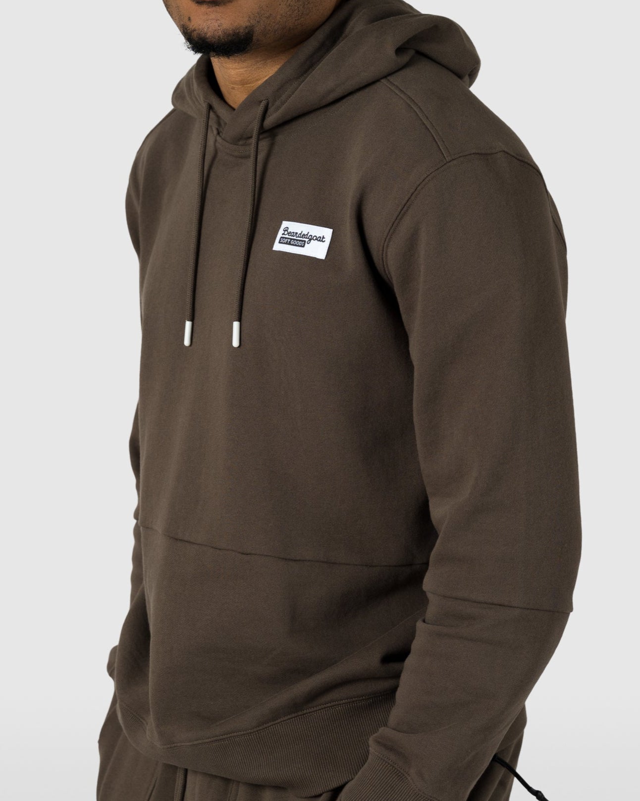 A young man with short curly hair stands against a light gray background wearing the BEARDEDGOAT Mens Drift Hoodie with a front pocket and matching brown sweatpants—an ideal lightweight outfit for spring to summer.