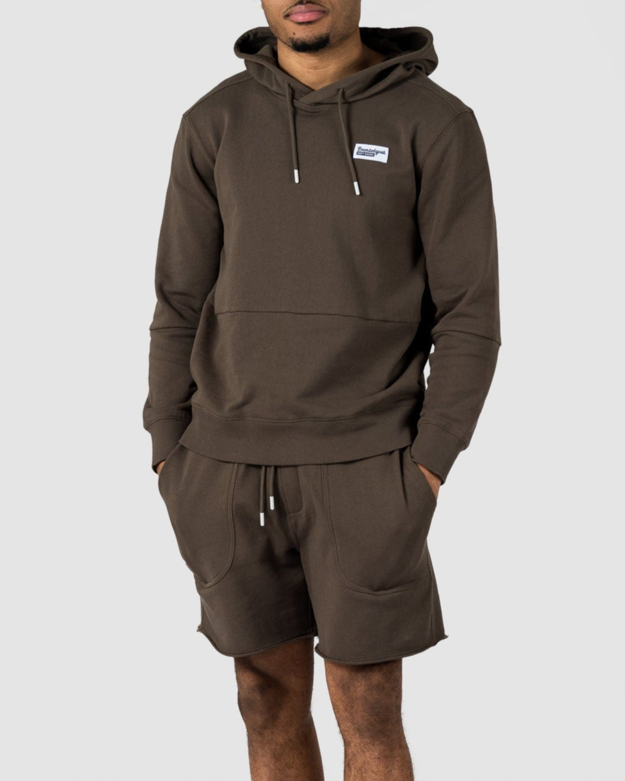 A man stands against a light gray background wearing the BEARDEDGOAT Mens Drift Hoodie in dark olive green with matching shorts, a small white patch, and white sneakers—an ideal look for transitioning from spring to summer.