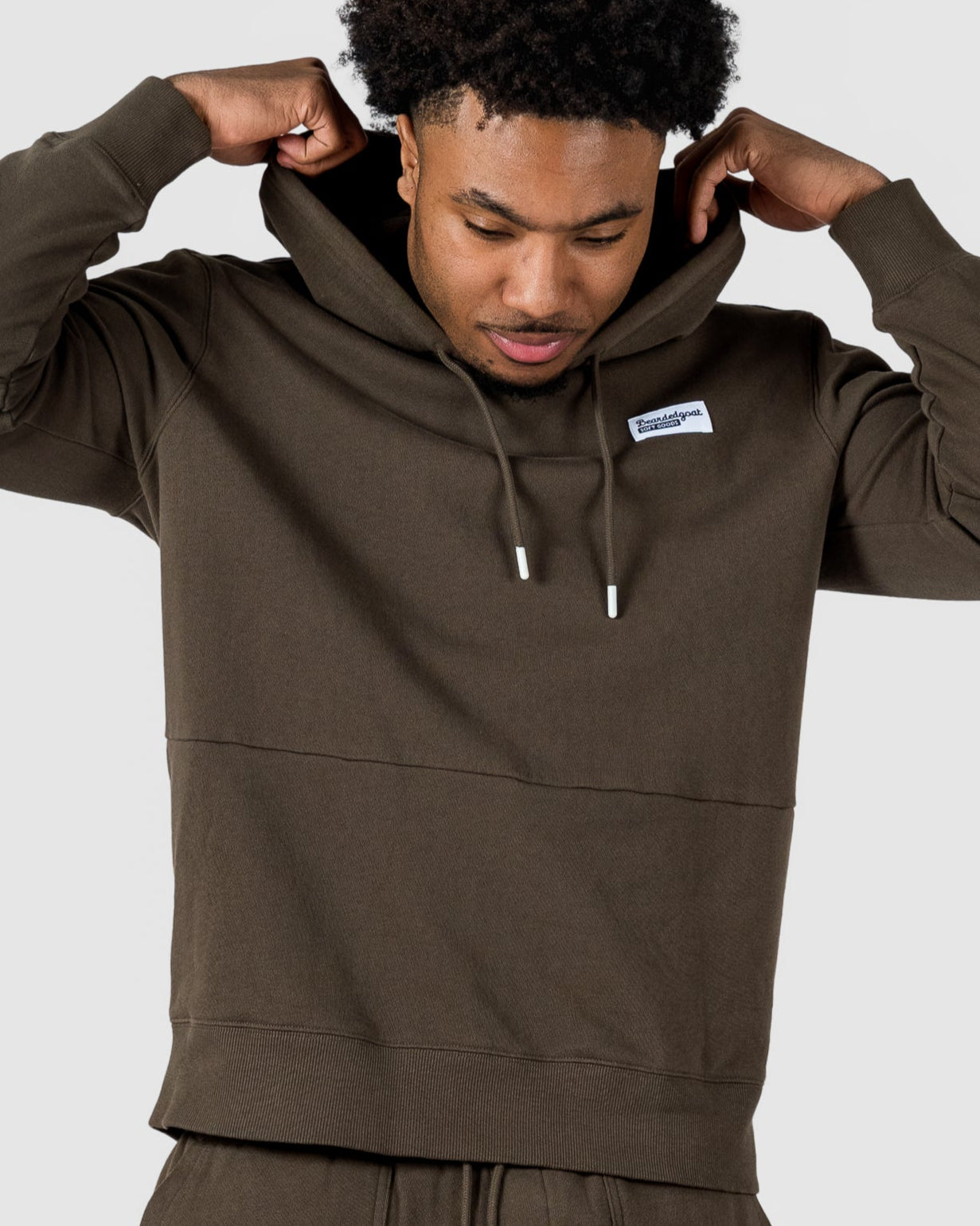 A man adjusts the hood of his BEARDEDGOAT Mens Drift Hoodie in dark olive green with matching shorts. Made from soft terry cloth, this lightweight outfit is ideal for spring to summer, shown against a plain light gray background.