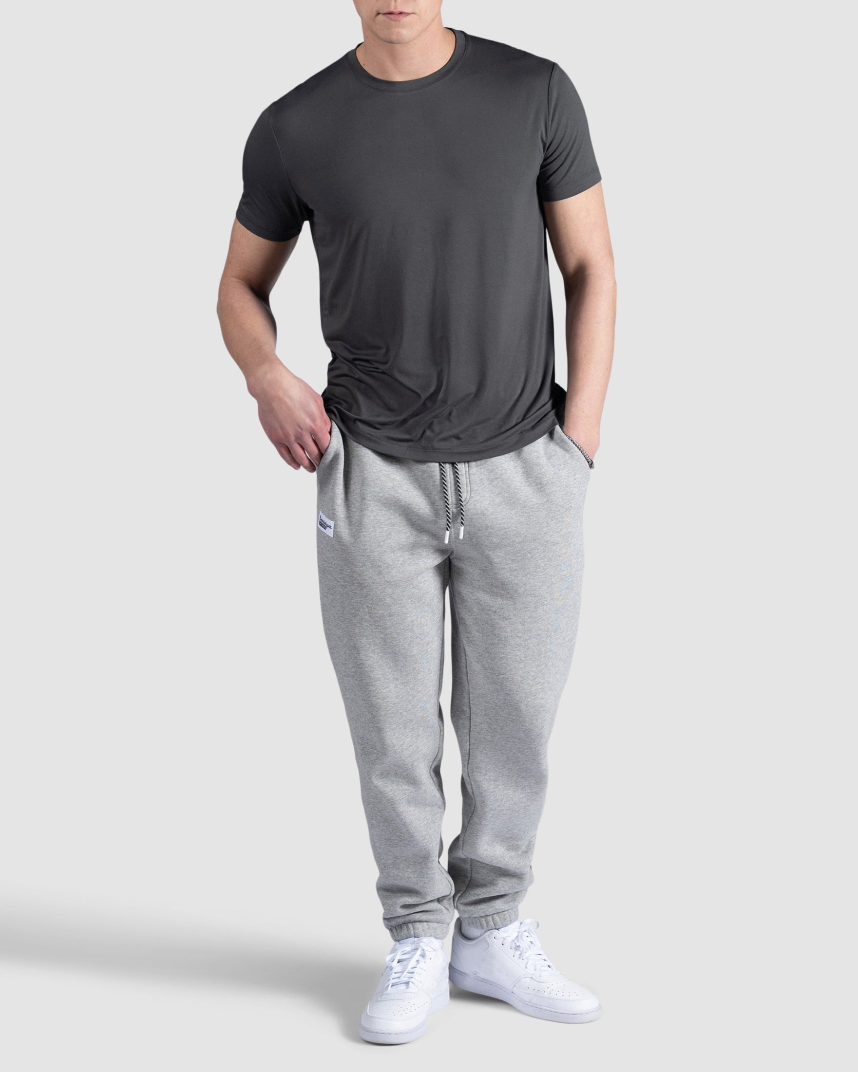 A man stands against a plain gray background, wearing the BEARDEDGOAT Mens EverZen Short Sleeve in dark gray, light gray sweatpants with a drawstring, and white sneakers. One hand is in his pocket while the other rests by his side.