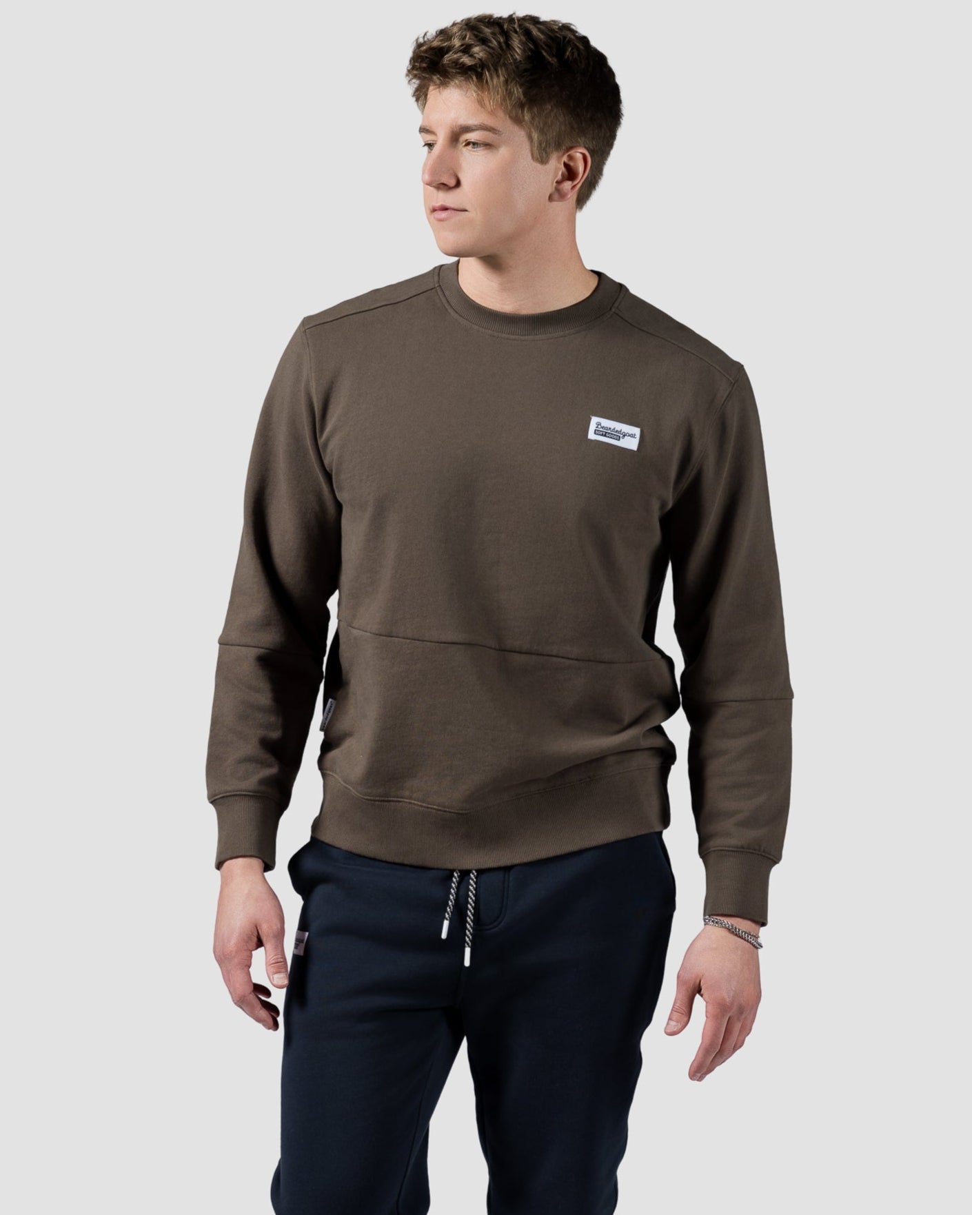 A man stands against a light background wearing the BEARDEDGOAT Mens Drift Crew sweatshirt, dark jogger pants, and white sneakers with blue and brown details. He looks to his left, embodying modern style and timeless charm.