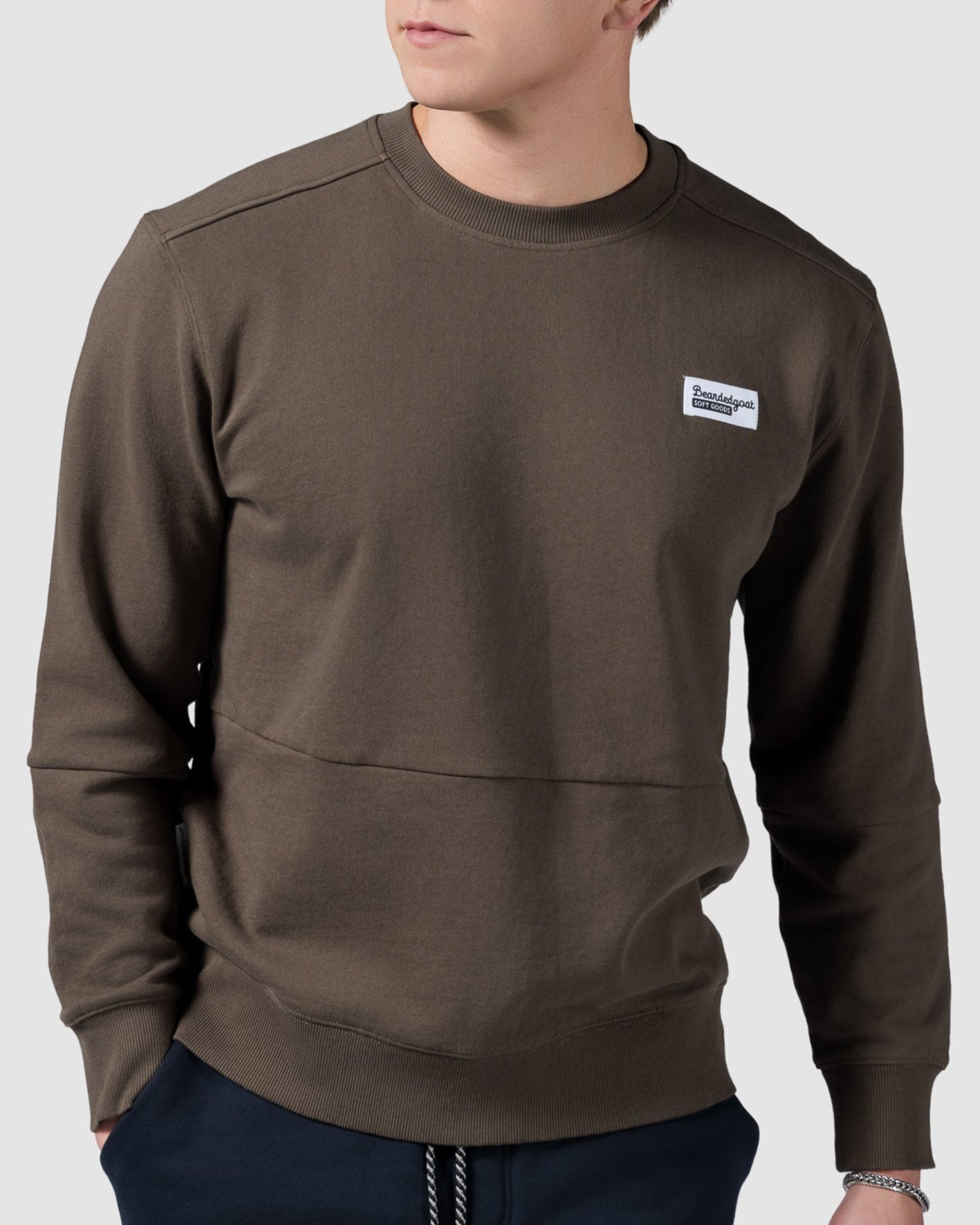 A young man with short brown hair wears the BEARDEDGOAT Mens Drift Crew sweatshirt and navy pants, hands in pockets, looking to the side—an easygoing, modern-meets-timeless look against a plain light background.