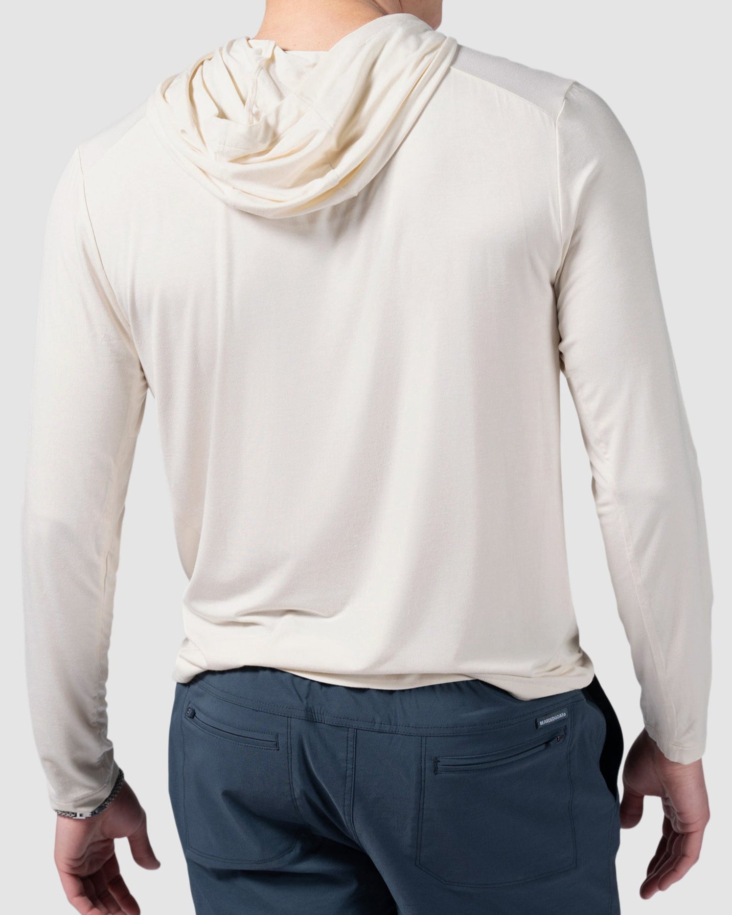 A man, seen from behind, wears a light beige BEARDEDGOAT Mens EverZen Hoodie crafted from natural bamboo fibers with blue pants against a plain light gray background.