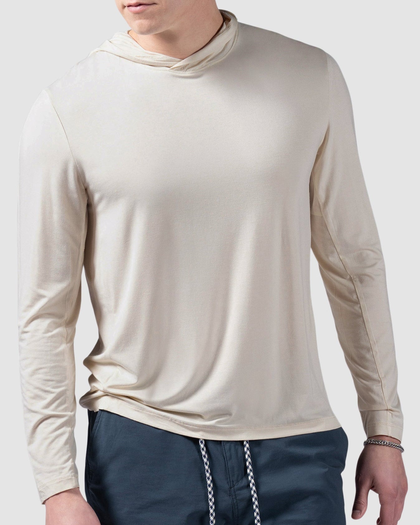 A young man in a beige BEARDEDGOAT Mens EverZen Hoodie, crafted from moisture-wicking bamboo fibers, and dark blue shorts with a white drawstring stands against a plain light gray background, looking to his left.