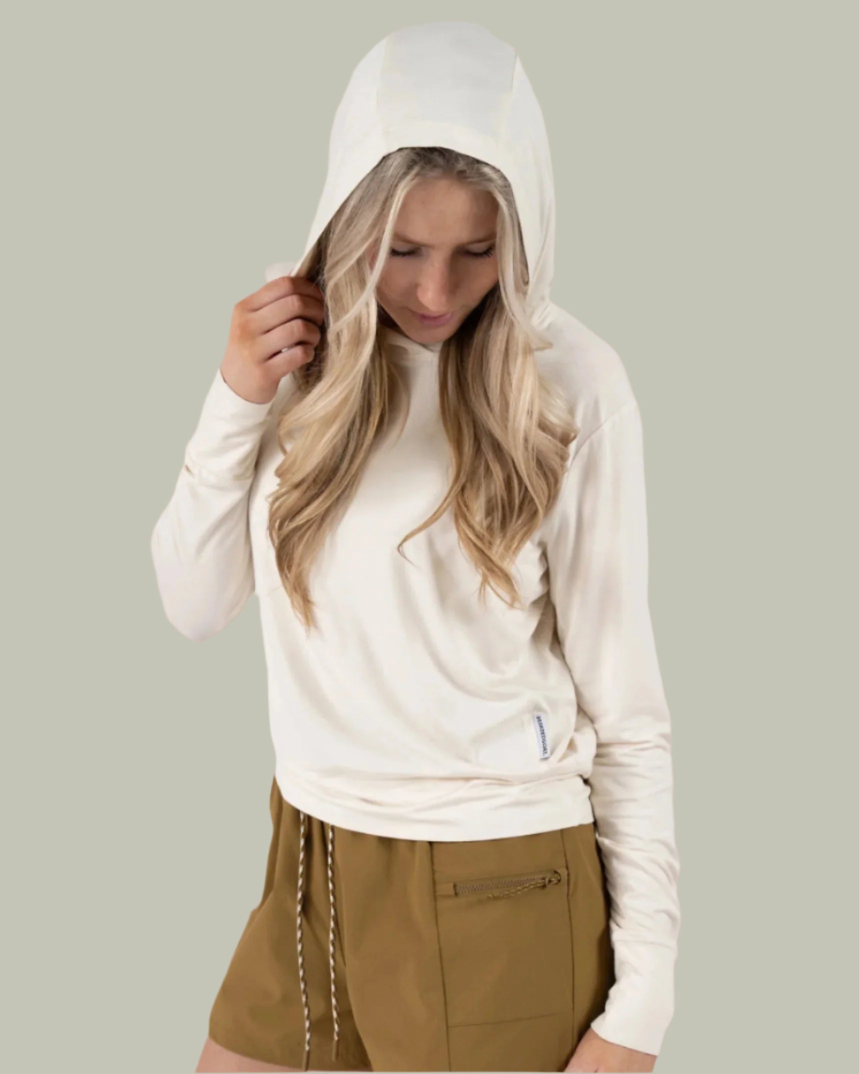 A woman stands against a light gray background, wearing the BEARDEDGOAT Womens EverZen Hoodie in cream and olive green drawstring shorts, looking down with one hand holding her hood.