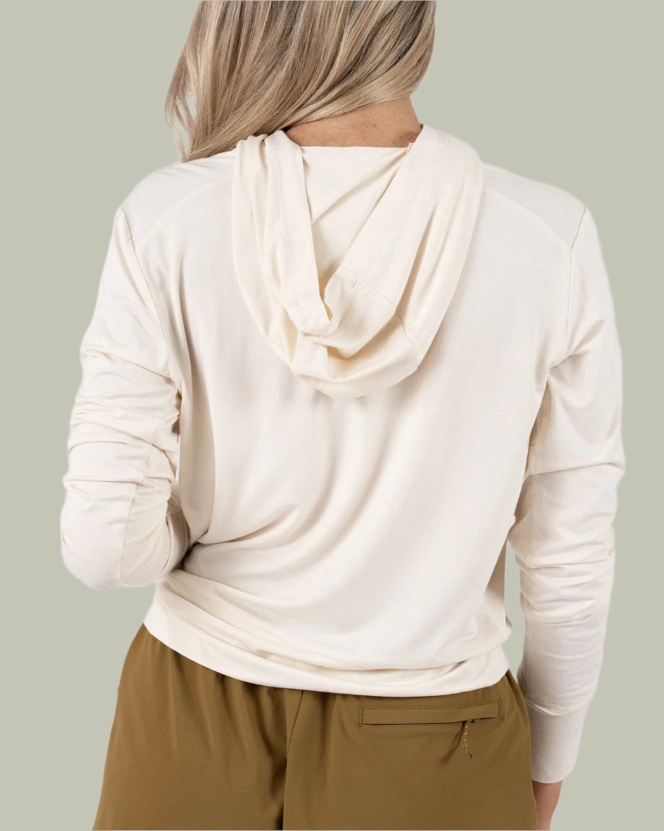 A woman with long blonde hair, seen from behind, wears the BEARDEDGOAT Womens EverZen Hoodie in light beige and olive green shorts against a plain light gray background. The hoodie is made from moisture-wicking bamboo fibers.