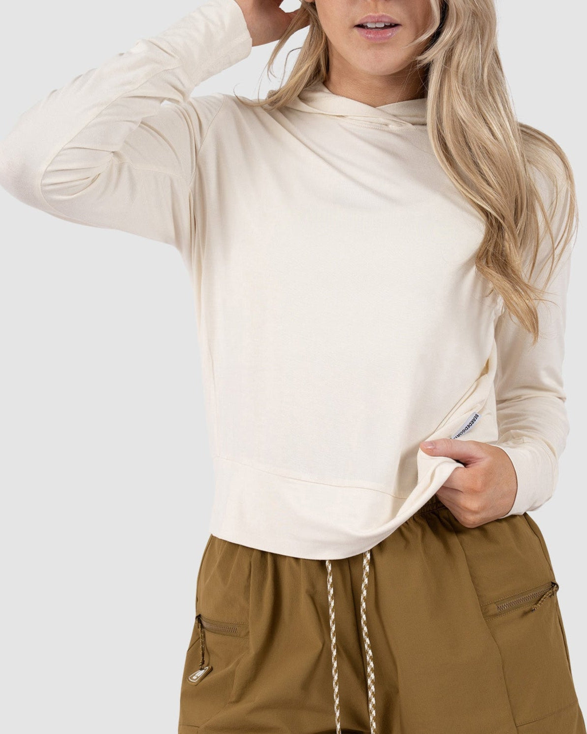 A woman with long blonde hair models the BEARDEDGOAT Womens EverZen Hoodie in cream, paired with brown zipper-pocket shorts, standing against a light gray background. She touches her head and holds her hoodie.