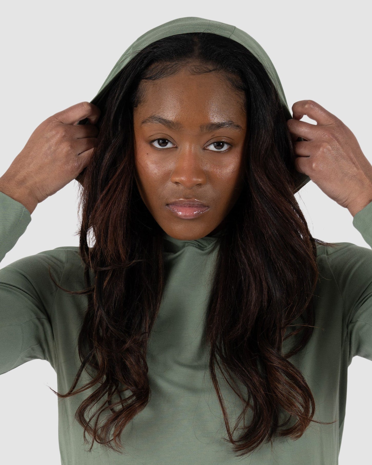 A woman with long dark hair wears the BEARDEDGOAT Womens EverZen Hoodie in sage green, crafted from soft bamboo fibers. She holds the hood with both hands and looks directly at the camera against a light gray background.