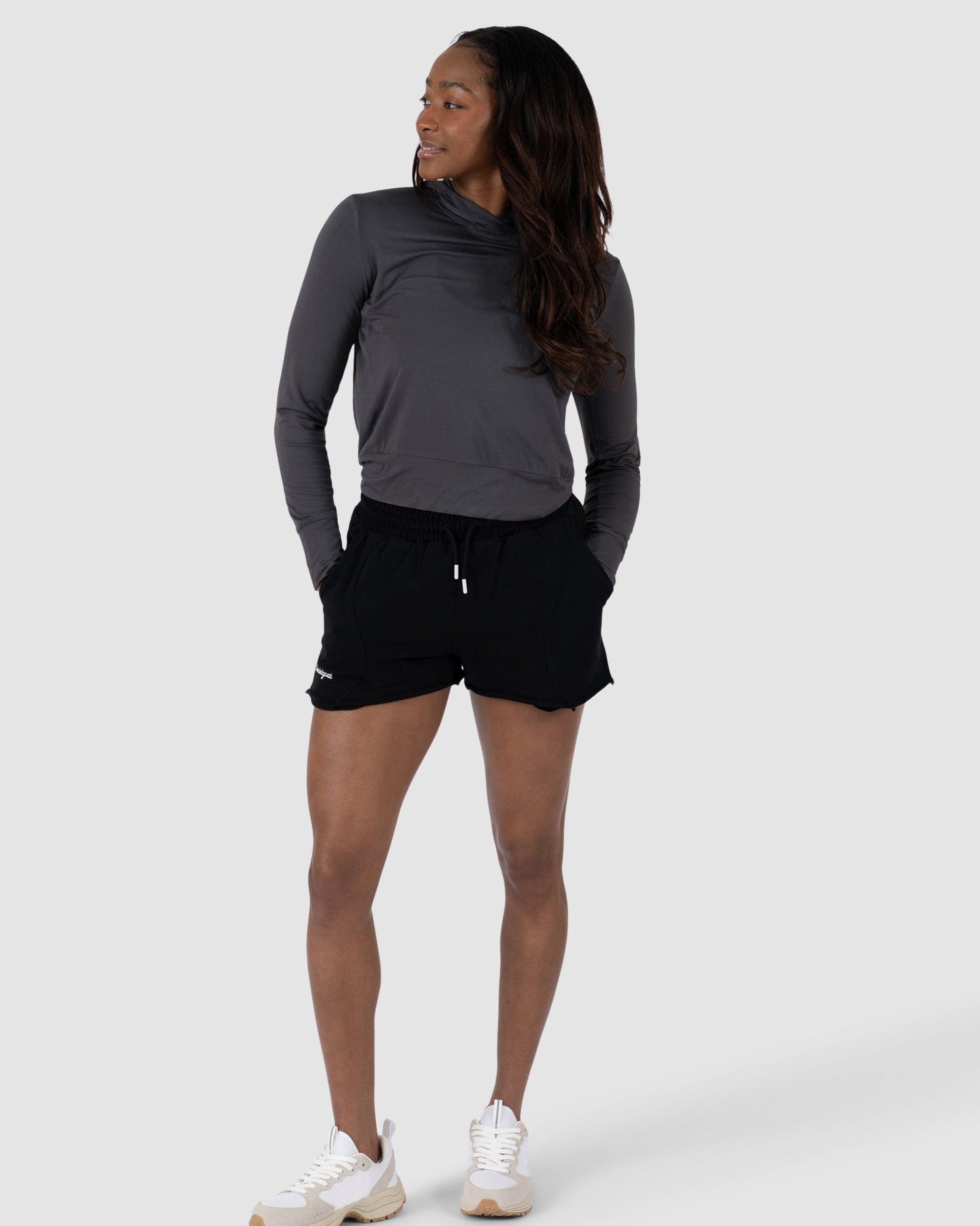 A woman in the BEARDEDGOAT Womens EverZen Hoodie, gray and moisture-wicking, stands with hands in her pockets. She pairs it with black shorts and white sneakers, glancing left against a plain light gray background.