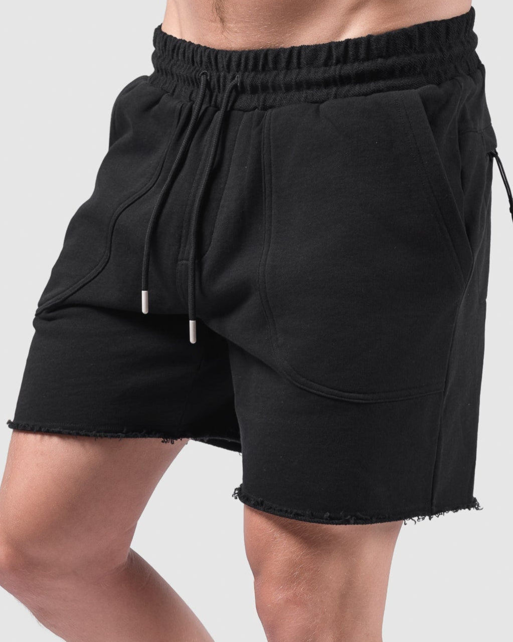 A person in BEARDEDGOAT Mens Drift Short in black, made from lightweight terry with frayed hems, pairs the shorts with white socks and sneakers. The face is not visible, and the background is light gray.
