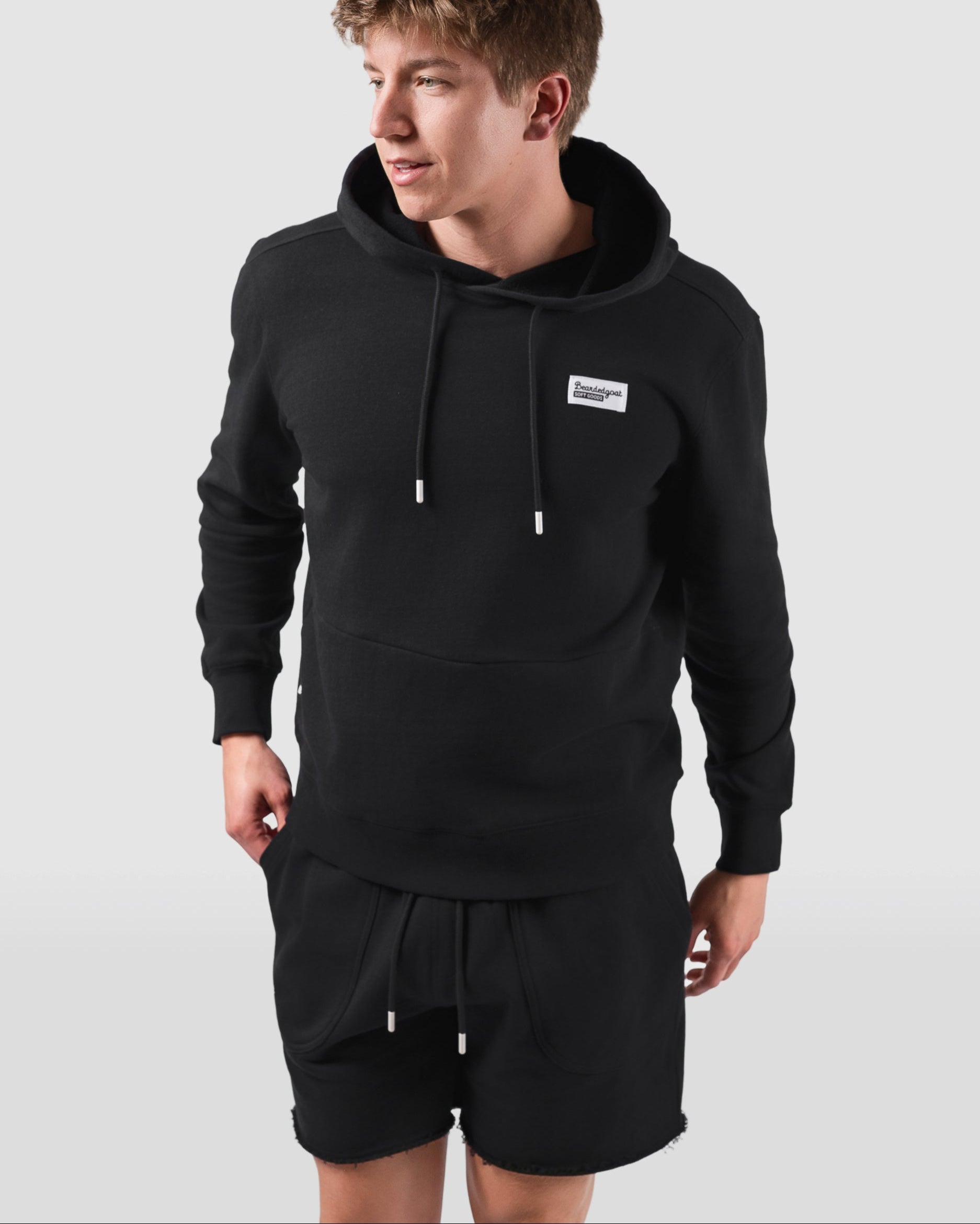A young man stands against a light gray background, looking to the side. He wears a black hoodie and BEARDEDGOAT Mens Drift Short, crafted from lightweight terry fabric, with his hands resting by his sides.