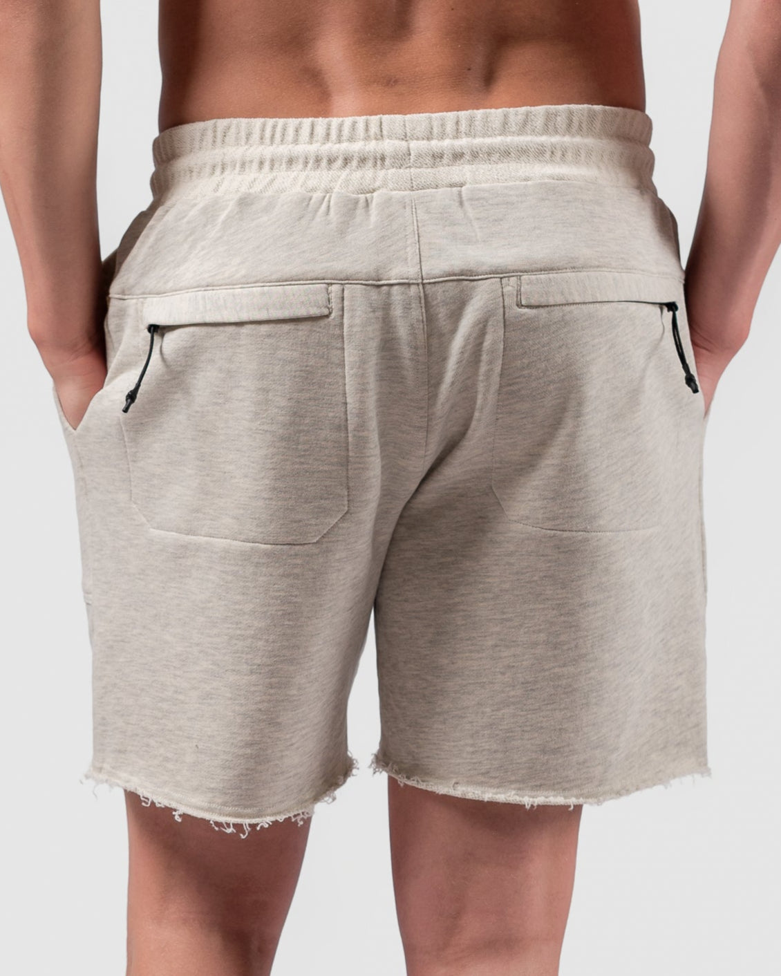A person stands facing away against a plain white background, wearing BEARDEDGOAT Mens Drift Shorts with frayed hems, paired with white socks and sneakers. Their hands are tucked into their pockets.