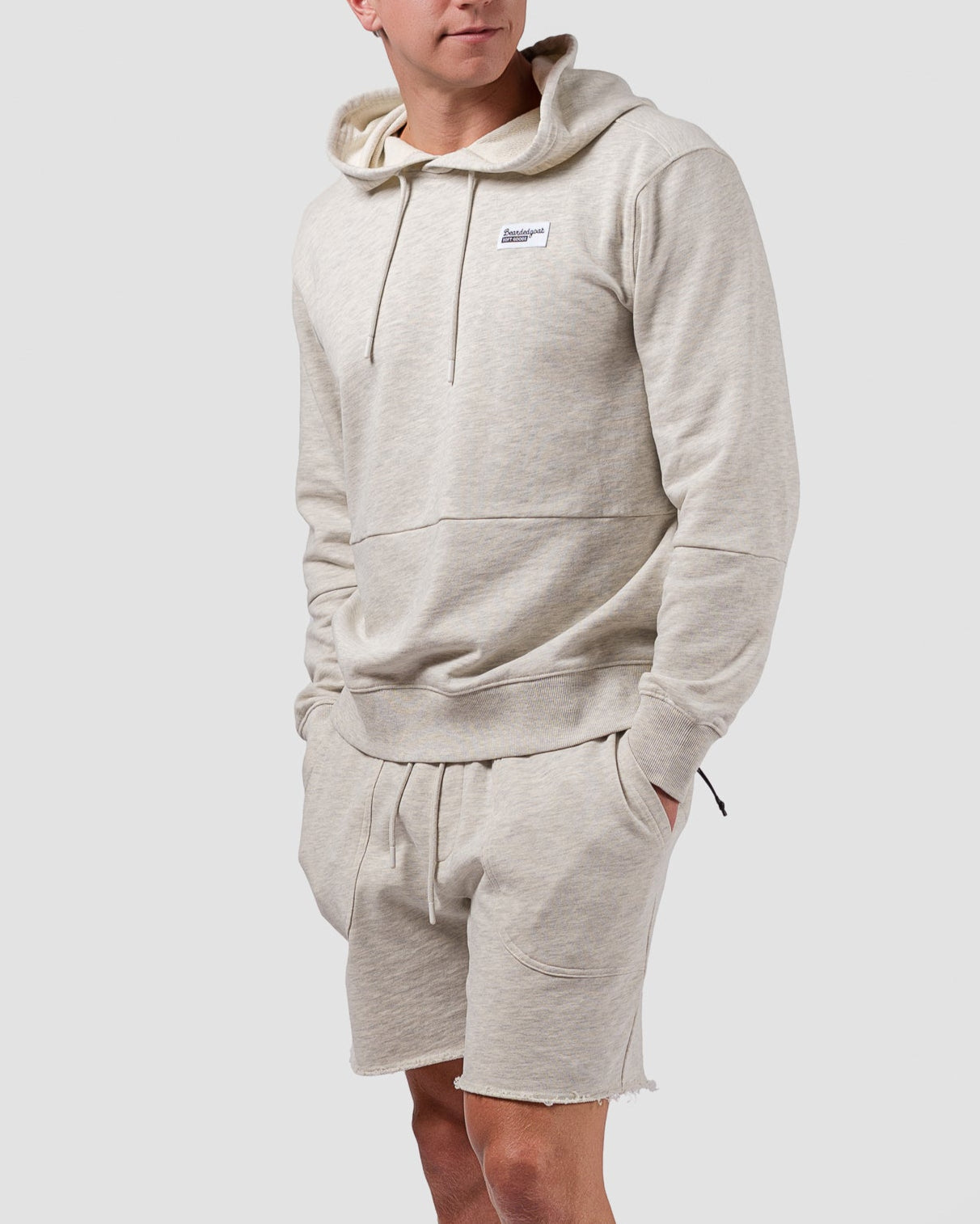 A young man stands against a plain background, wearing BEARDEDGOAT Mens Drift Short and a matching light beige hooded sweatshirt, paired with white crew socks and sneakers. His hands are in his pockets as he looks slightly to the side.