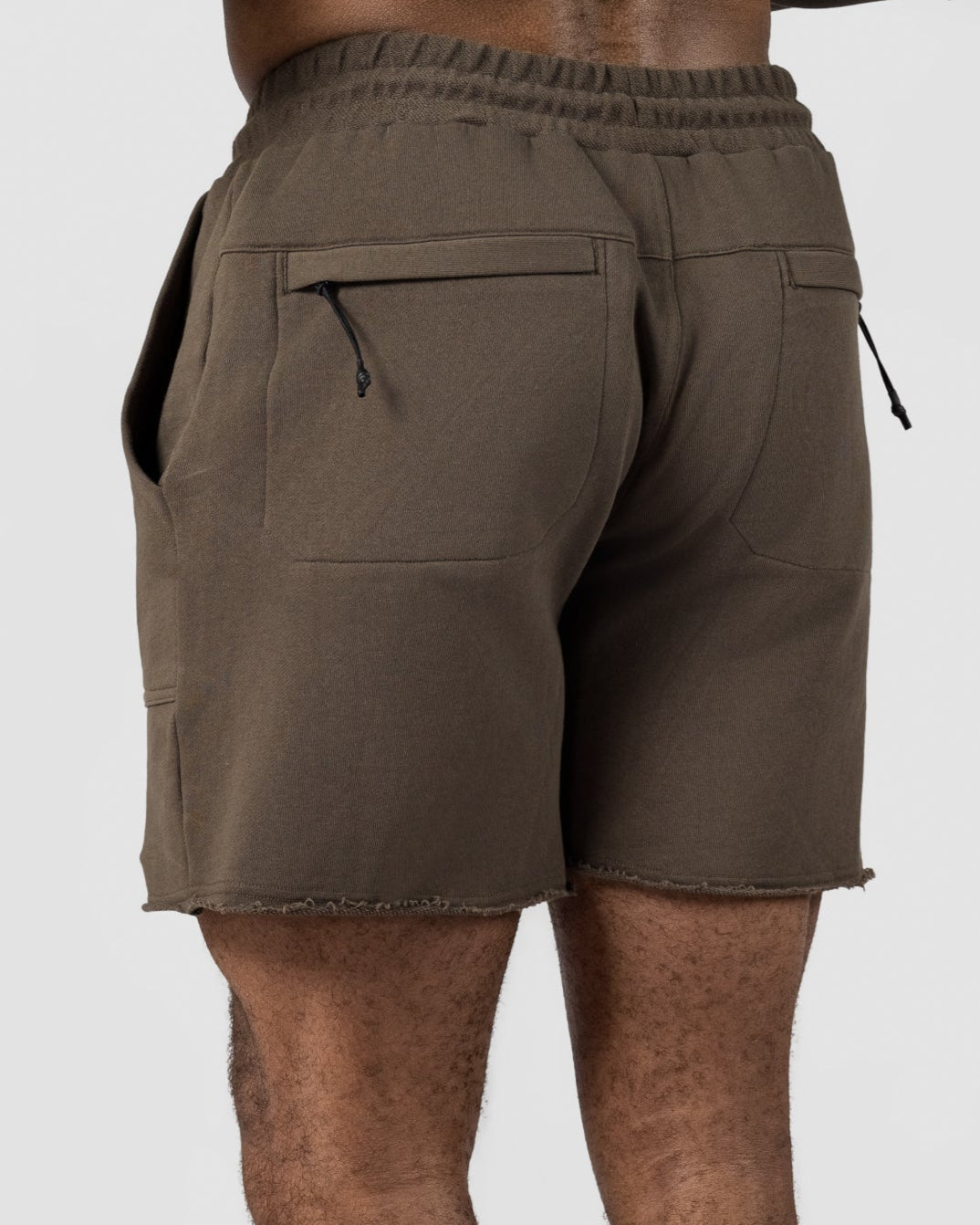 A person is seen from the back wearing olive green BEARDEDGOAT Mens Drift Shorts made from lightweight terry fabric, paired with white Nike socks and sneakers, against a plain light gray background.