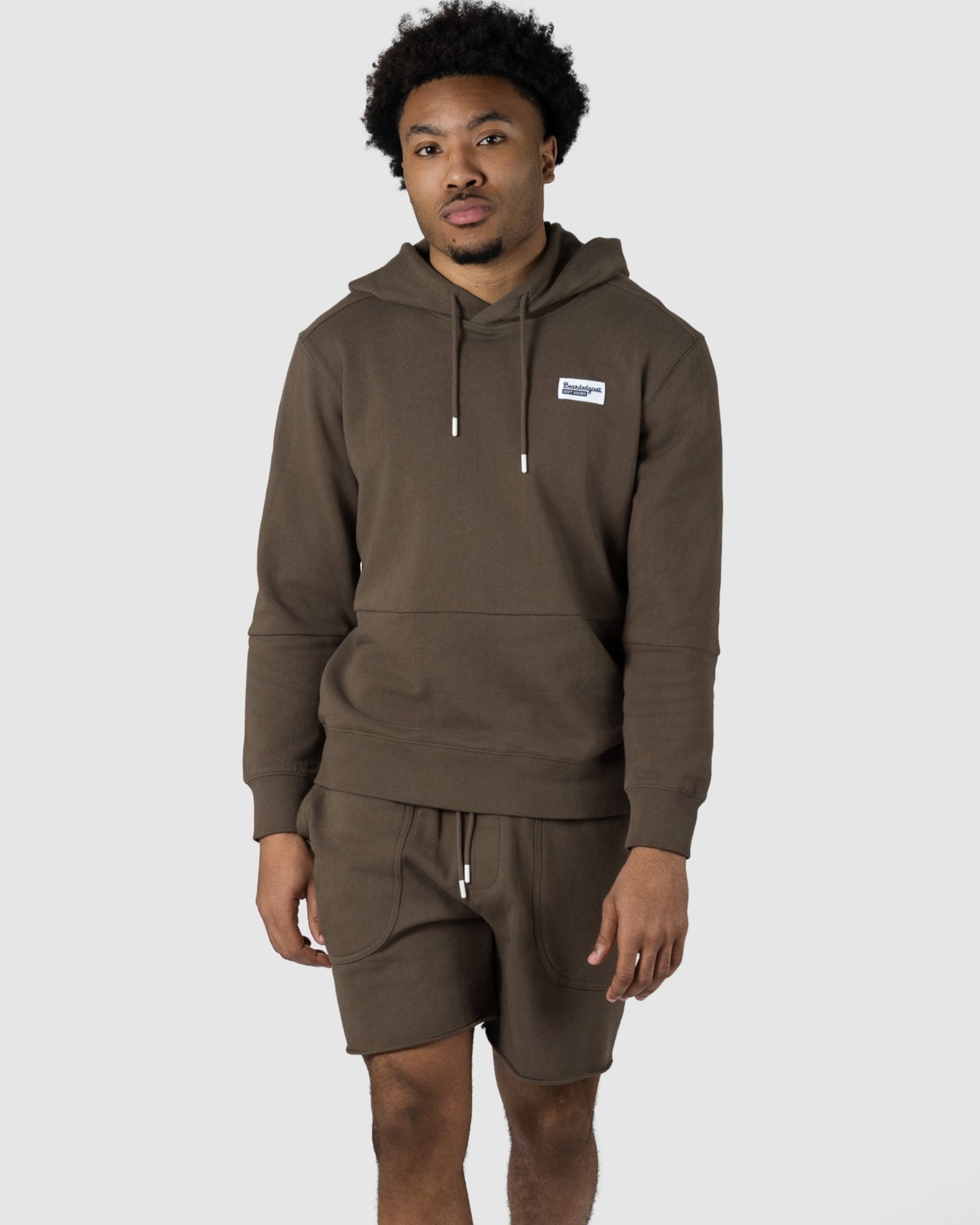 A man in an olive green BEARDEDGOAT Mens Drift Short and matching terry hoodie walks forward in white sneakers. His short curly hair complements the relaxed, lightweight look, set against a plain light gray background.