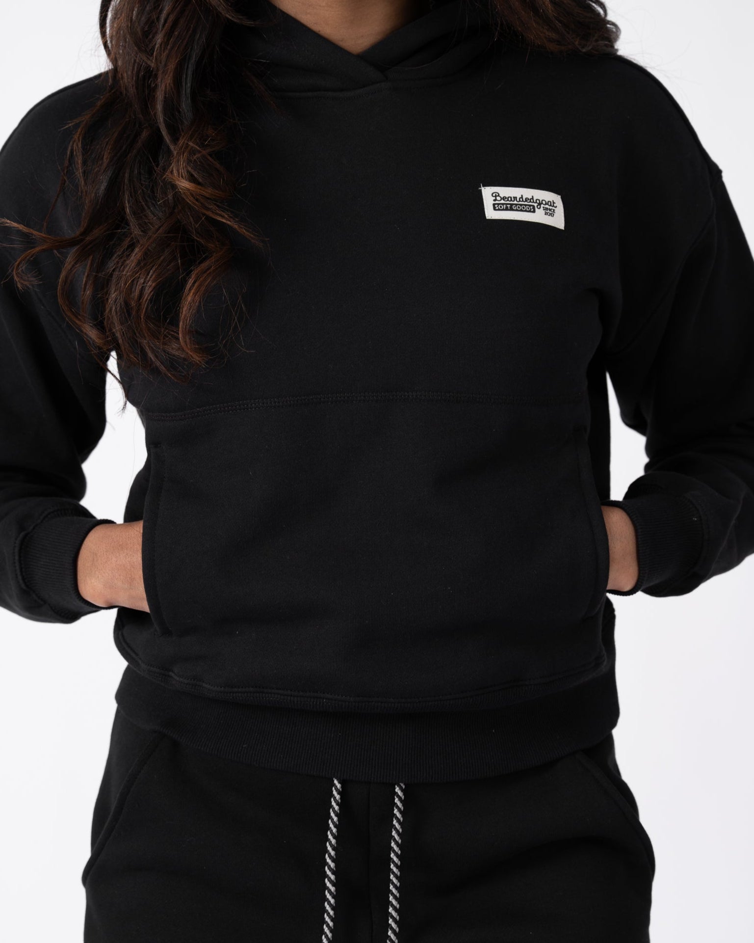 A woman wears the beardedgoat Womens Overnight Hoodie in black heavyweight cotton fleece, featuring a small rectangular patch, paired with matching black sweatpants and white drawstrings, hands in her hoodie pocket, against a plain white background.