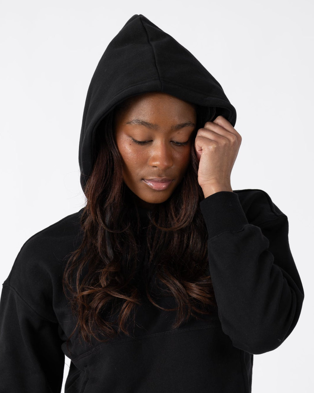 A woman wearing beardedgoat’s Womens Overnight Hoodie in black heavyweight cotton fleece with matching sweatpants stands against a plain white background, hood up, looking down as she touches the edge of her hood.