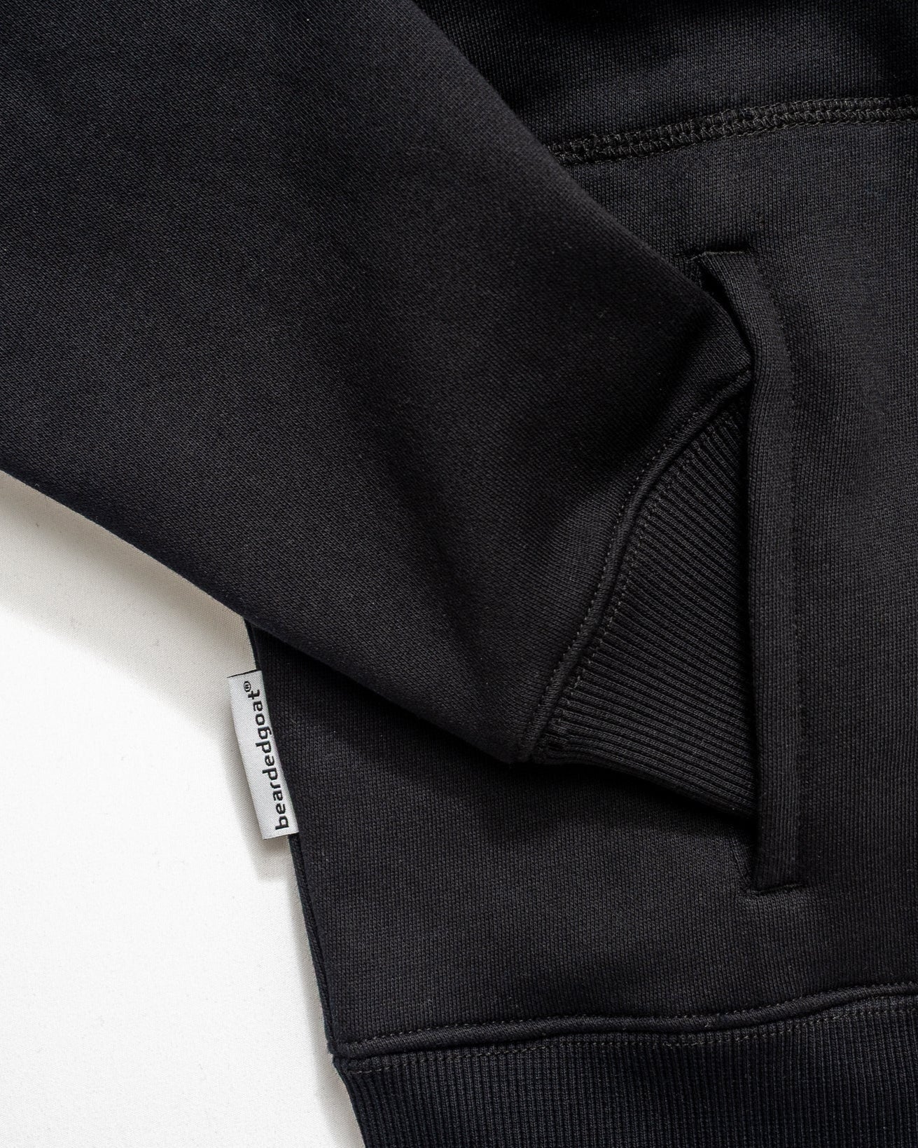 Close-up of the Womens Overnight Hoodie by beardedgoat in black heavyweight cotton fleece, featuring a front pocket, ribbed hem, and a small white branded tag on the seam, displayed on a white background.