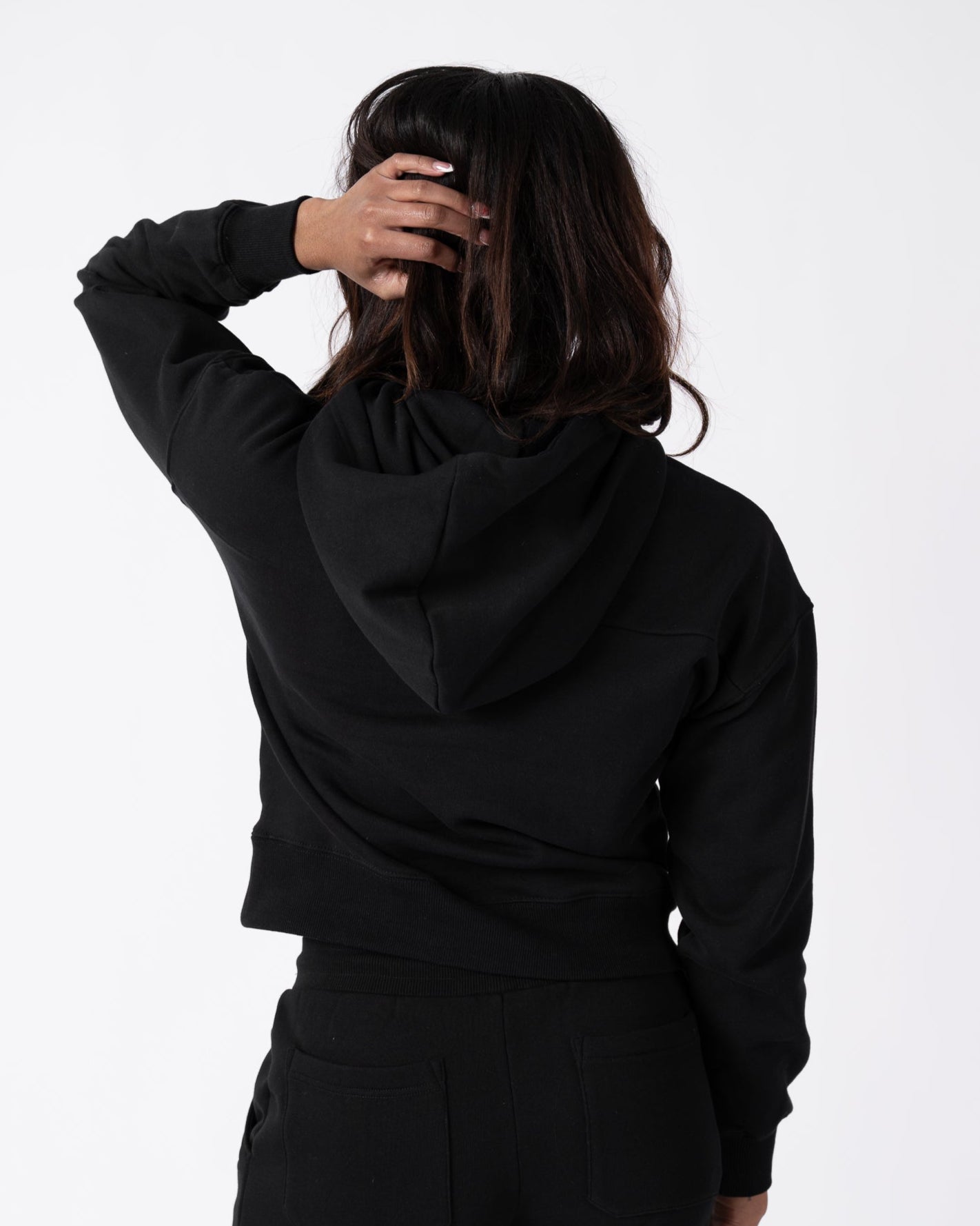 A woman wearing the beardedgoat Womens Overnight Hoodie in black heavyweight cotton fleece and black pants stands facing away, touching the back of her head against a plain white background.