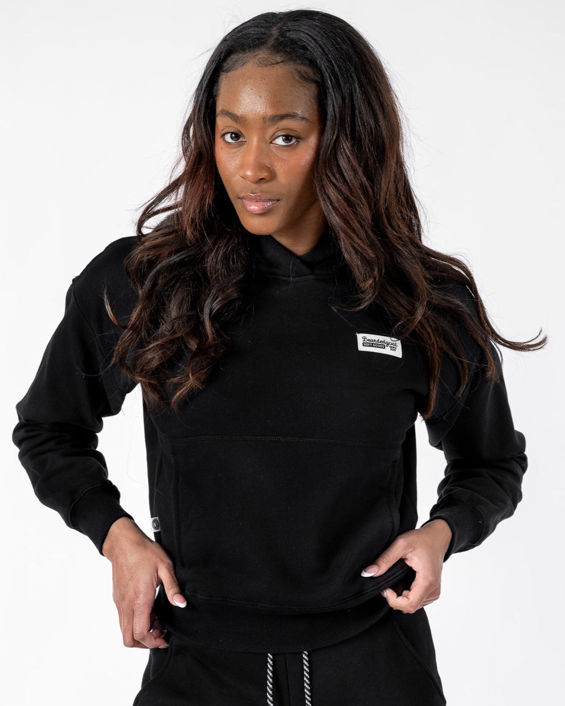 A woman with long dark hair stands against a plain white background, wearing the beardedgoat Womens Overnight Hoodie made from heavyweight cotton fleece and matching sweatpants, looking at the camera with a neutral expression.