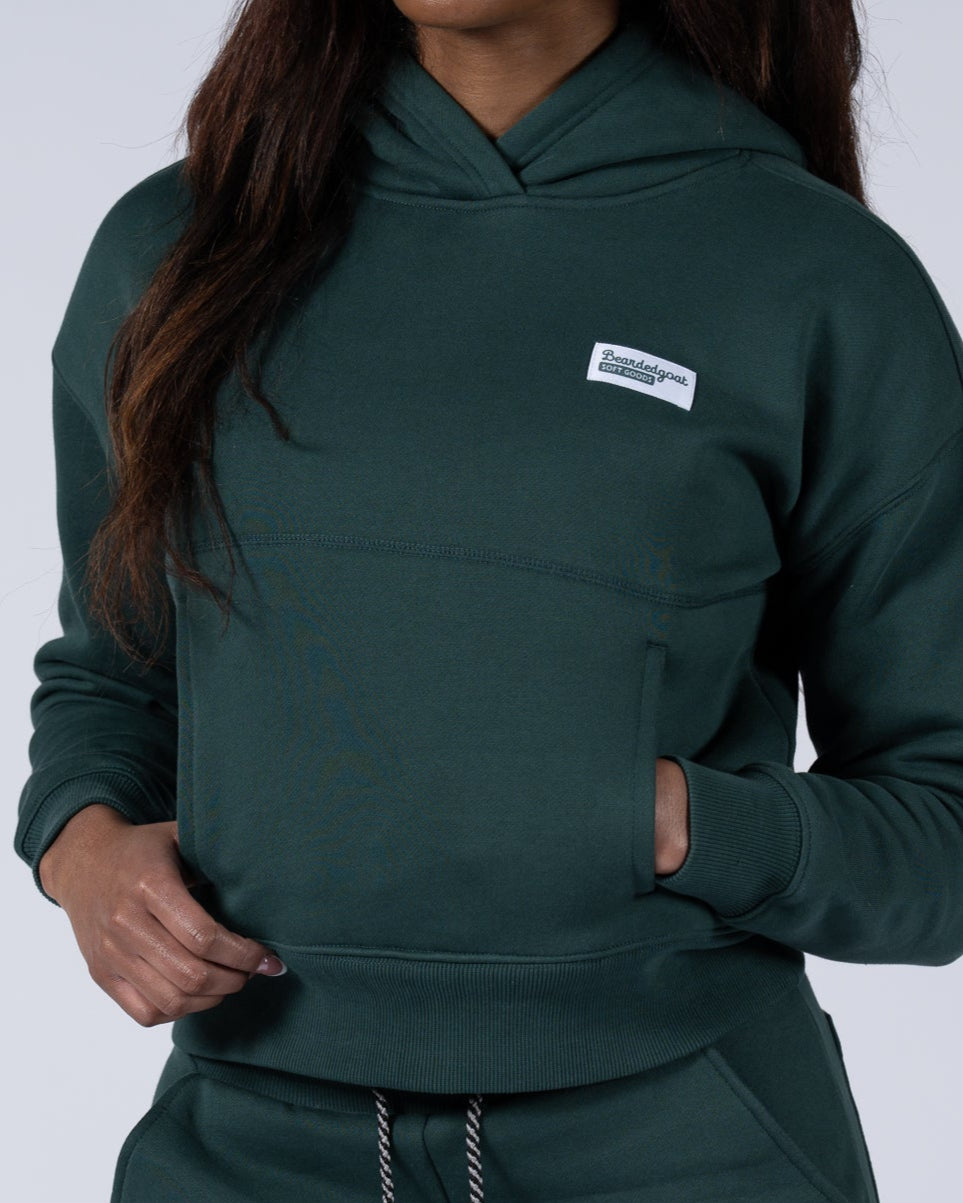 A BG woman with long dark hair wears the beardedgoat Womens Overnight Hoodie in dark green heavyweight cotton fleece, standing against a light gray background with her hands in her pockets.
