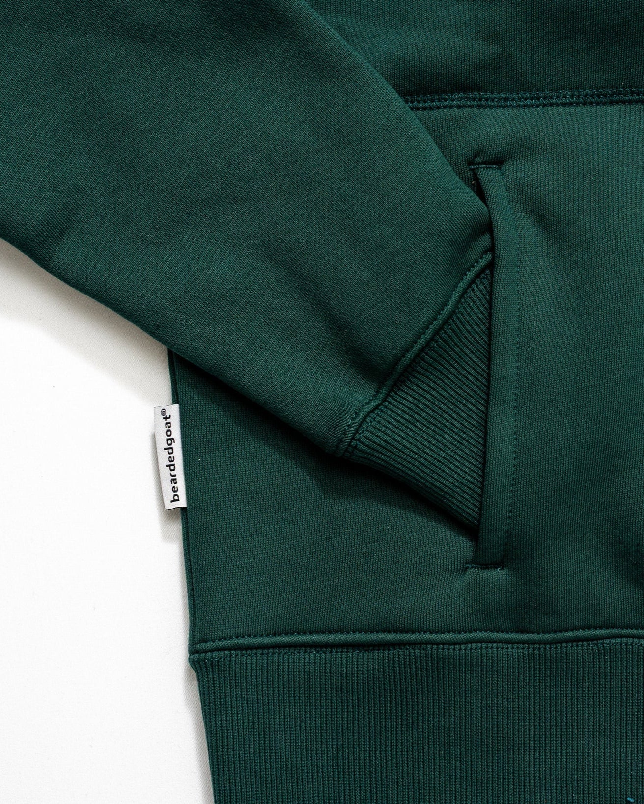 Close-up of the beardedgoat Womens Overnight Hoodie in dark green heavyweight cotton fleece, featuring a front pocket, ribbed cuff, and small white fabric tag with text on the side seam.