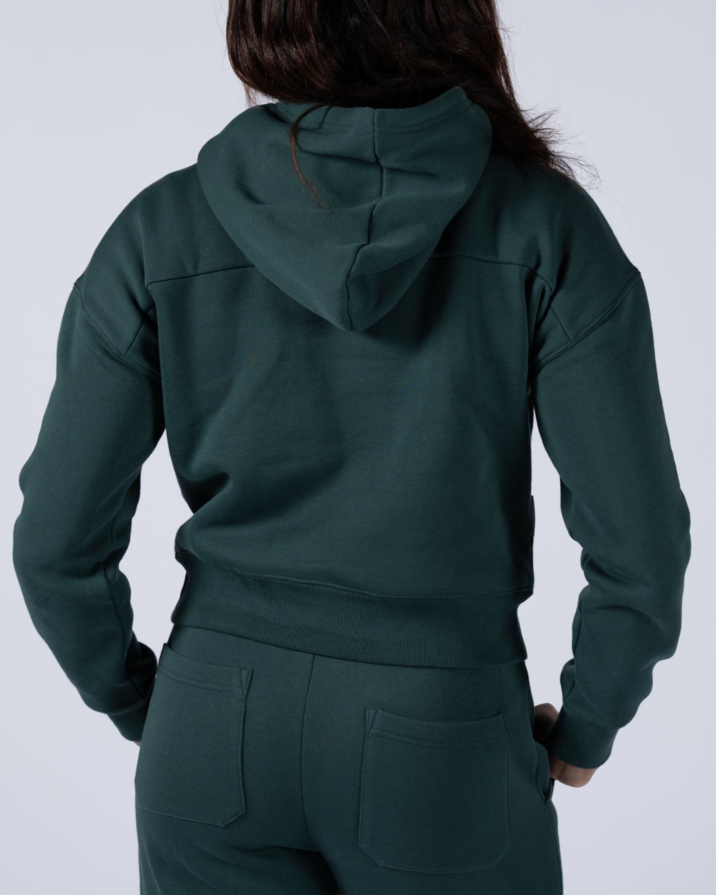 A woman with long dark hair faces away from the camera, wearing the beardedgoat Womens Overnight Hoodie and matching green pants against a plain light background.